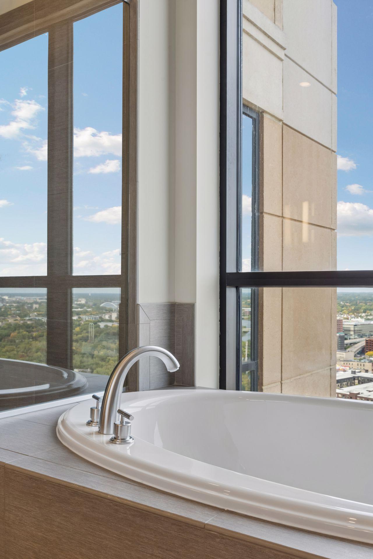 Soaking tub beside floor-to-ceiling windows with a city view.