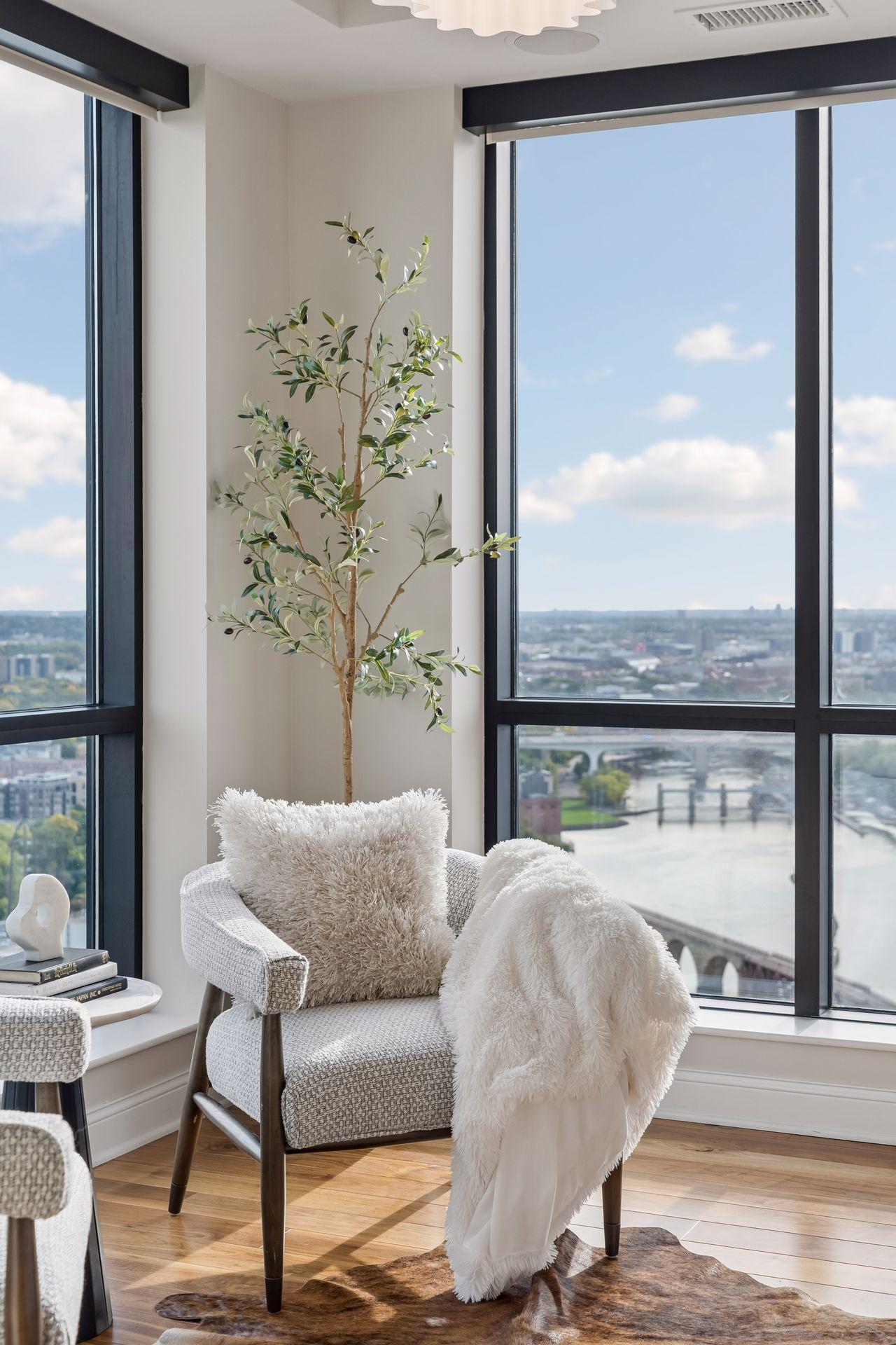 Staged condo sitting area with a white chair and olive tree beside windows overlooking the river and city.