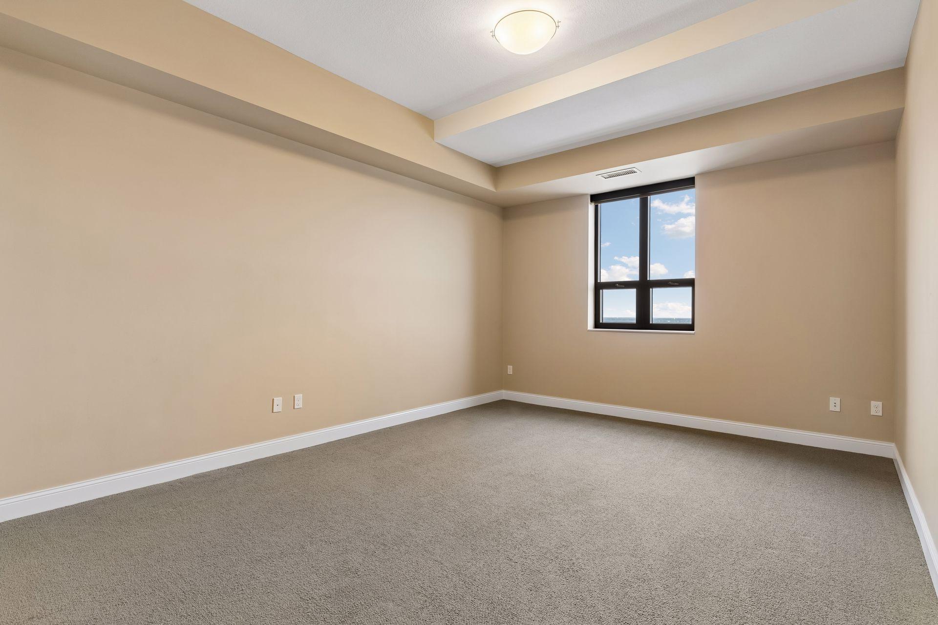 Empty carpeted room with a recessed ceiling and a window showing blue sky and water.
