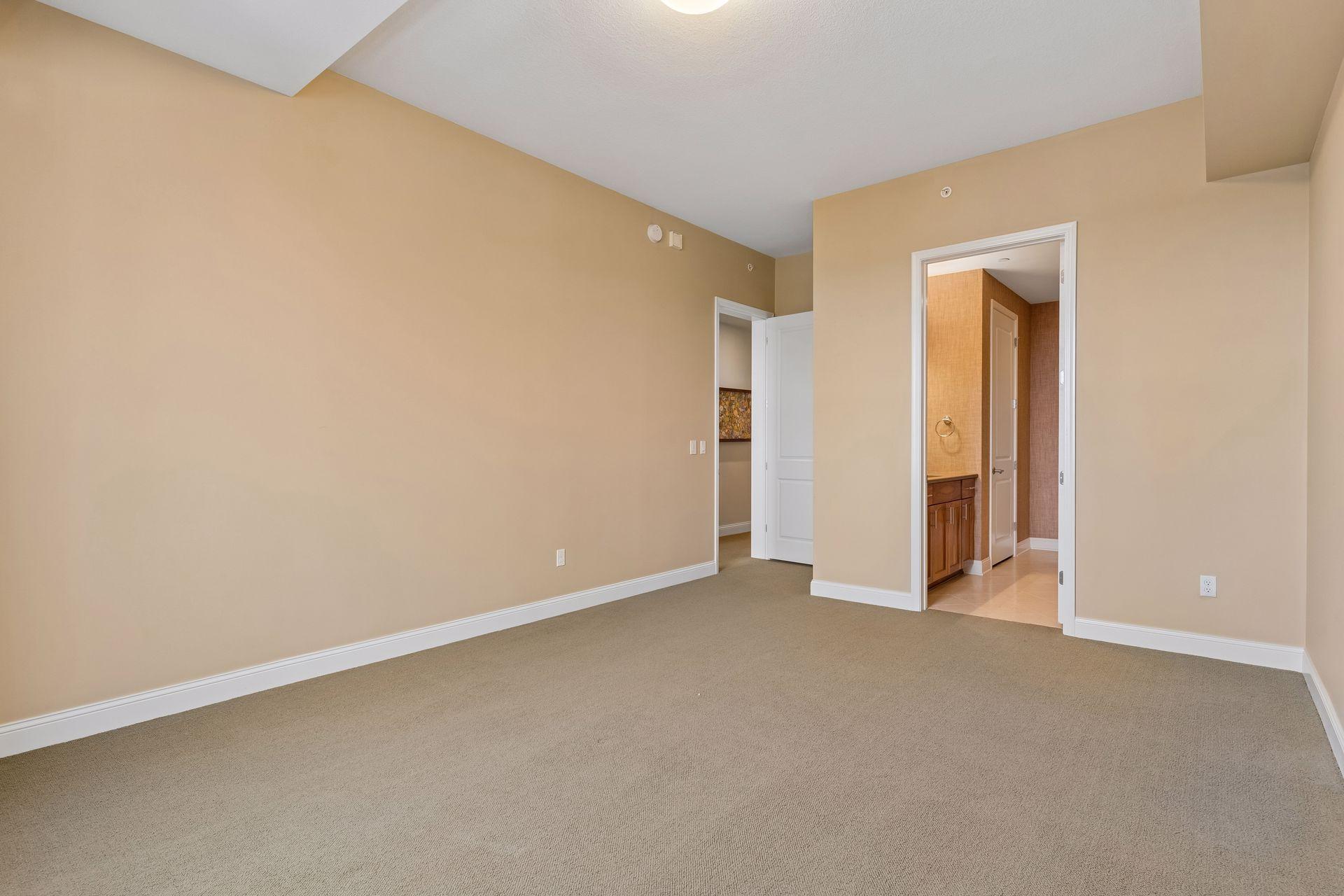Empty carpeted room with beige walls and an open doorway to a bathroom vanity.
