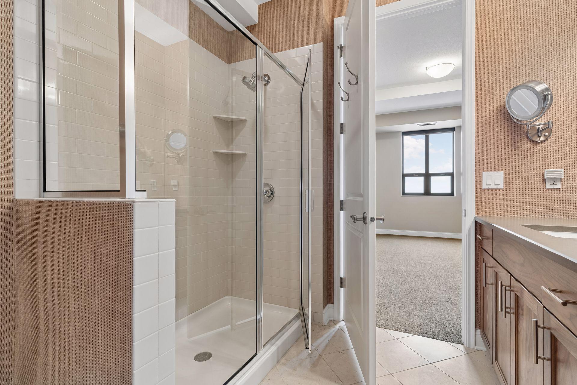 Condo bathroom with a glass-enclosed tiled shower and wood vanity, opening onto a carpeted room.