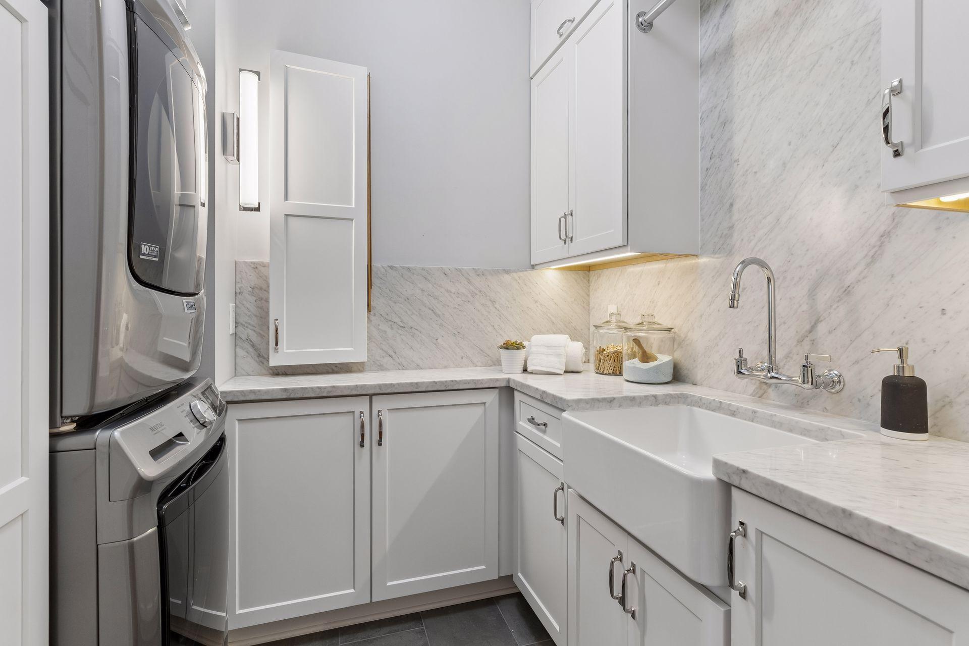 Laundry room with stacked washer and dryer, white cabinetry, marble surfaces, and a farmhouse sink.