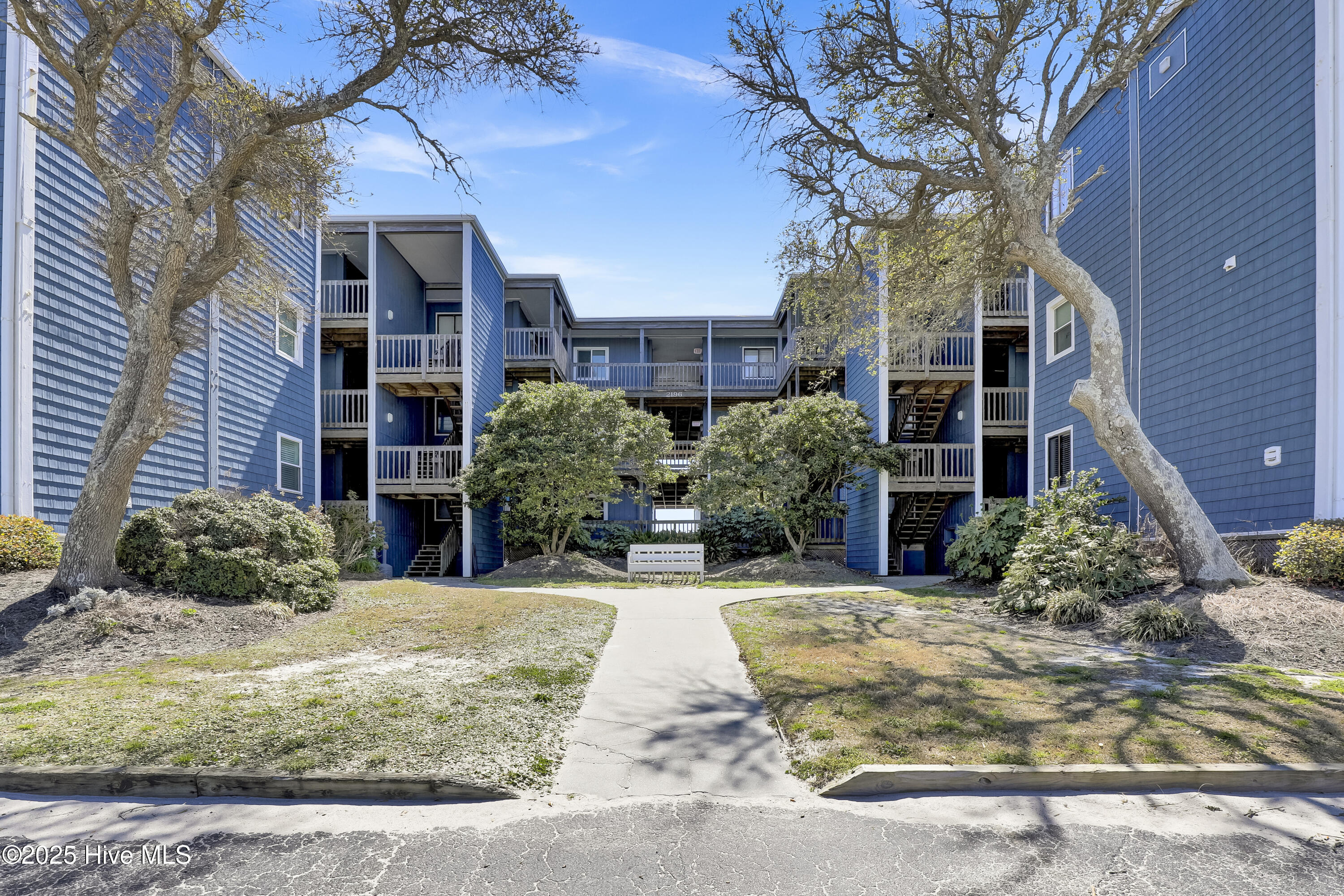 Blue condominium building with balconies and exterior stairs, framed by mature trees and a landscaped courtyard.