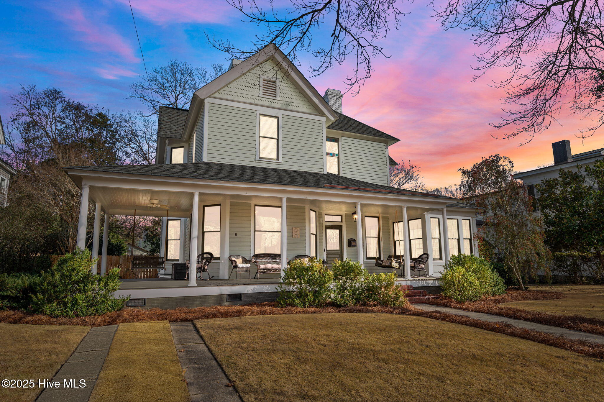 Two-story gray house with a long columned porch beneath a pink and orange dusk sky.