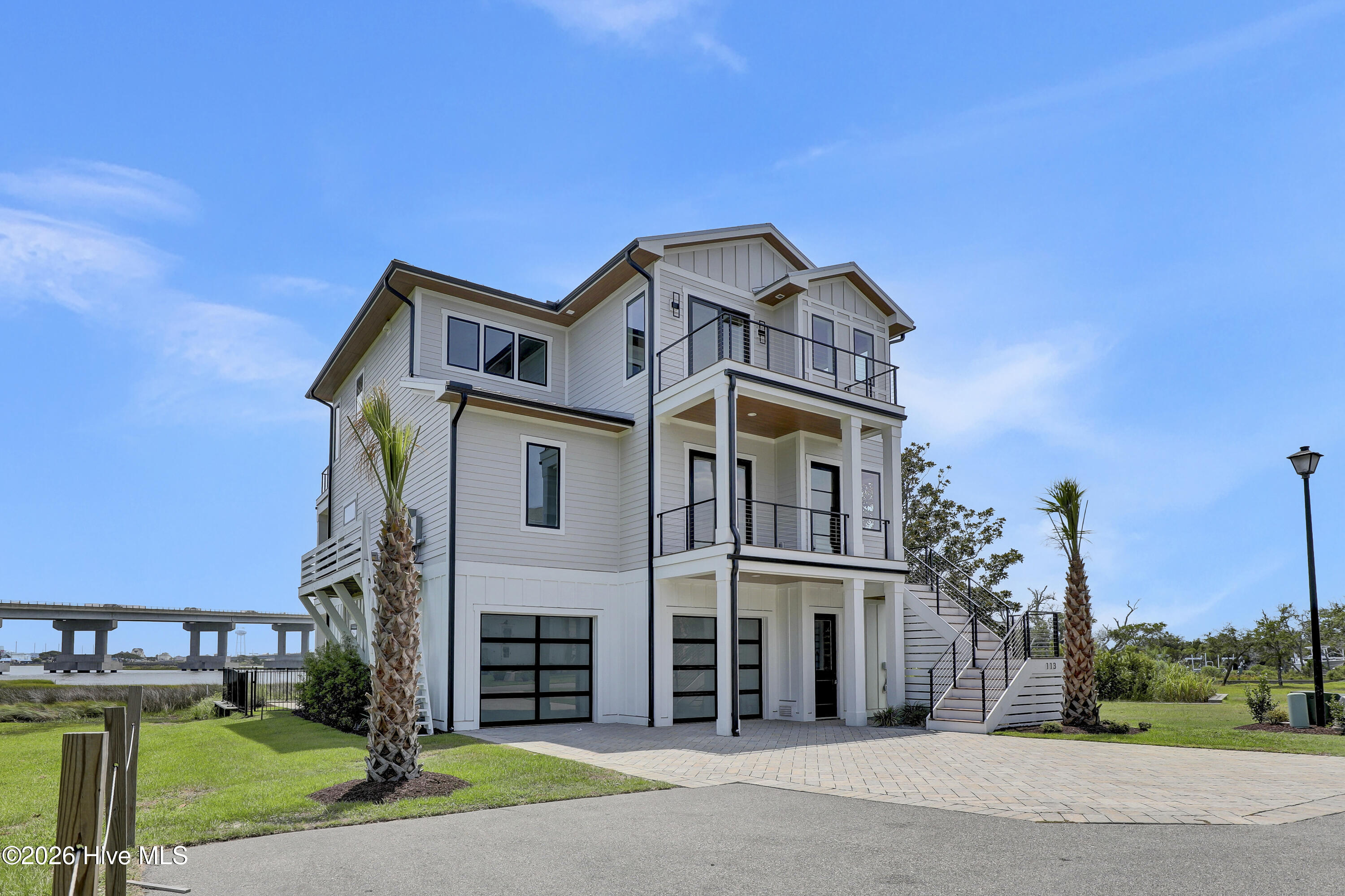 Modern white three-story waterfront home with balconies, exterior stairs, and a bridge in the background.
