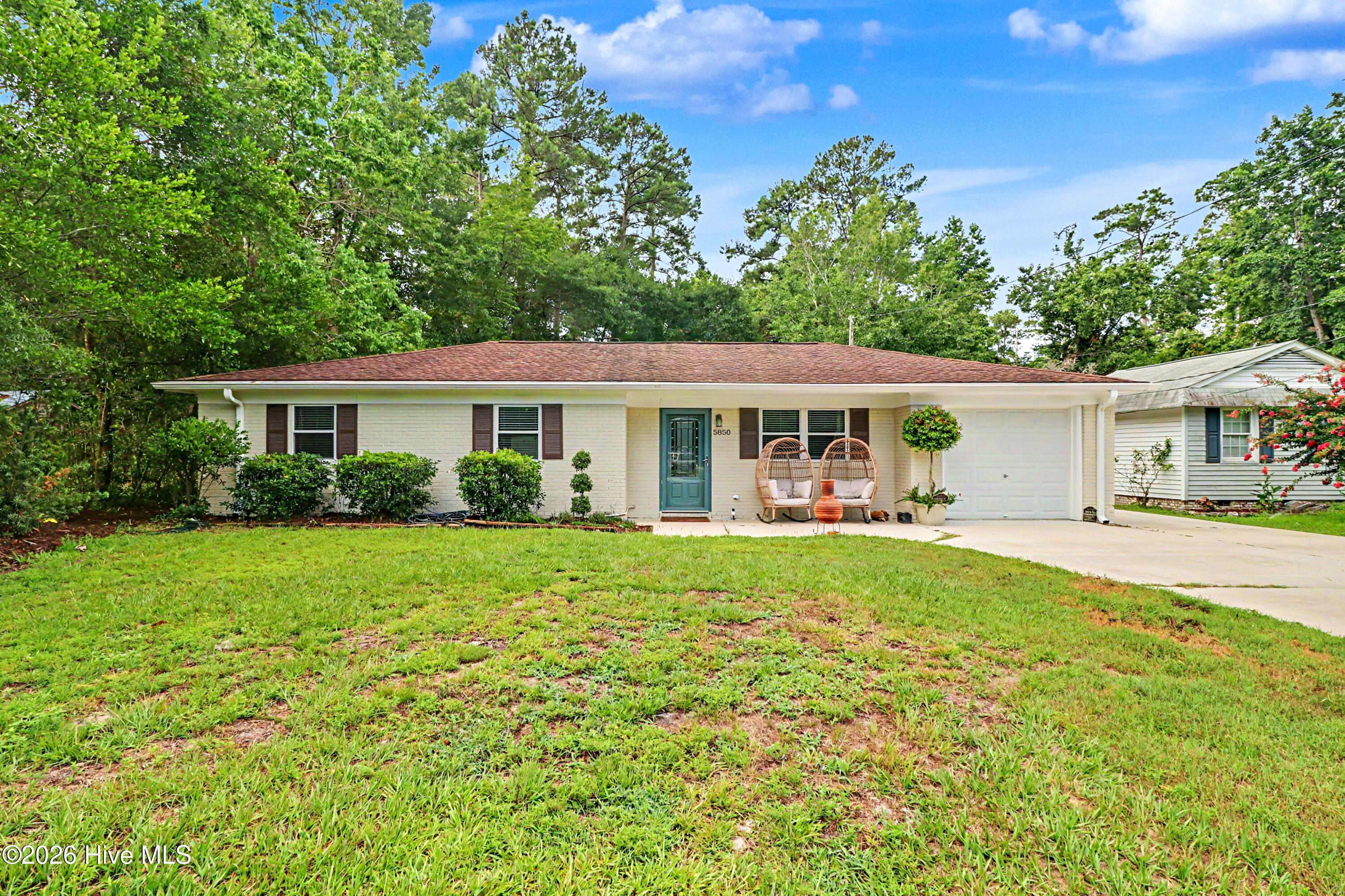 Single-story white brick home with a teal front door, attached garage, and wicker chairs at the entrance.