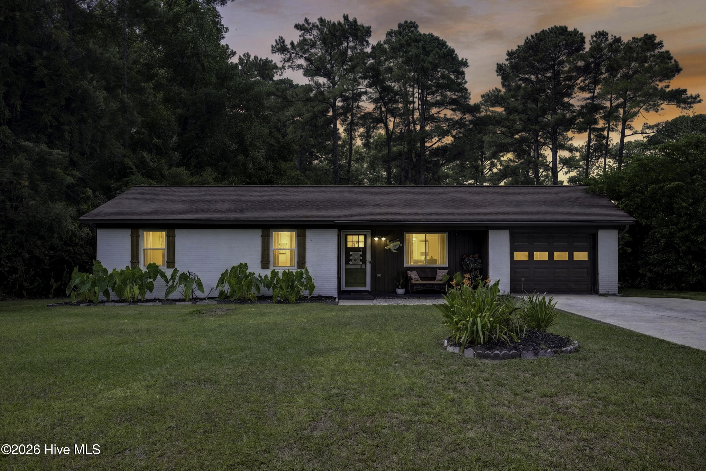 Single-story white ranch home with an attached garage, landscaped lawn, and wooded backdrop at dusk.