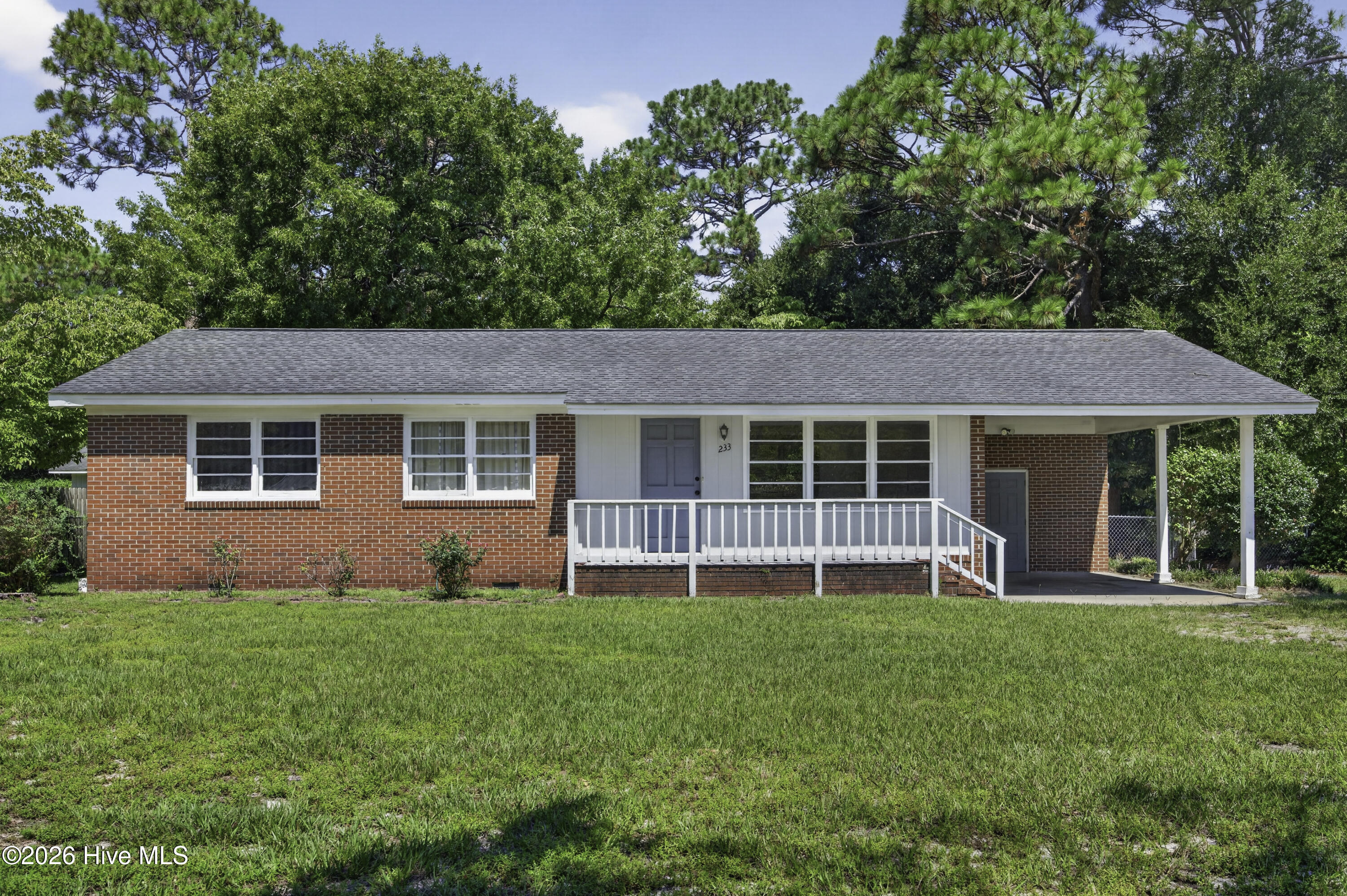 Single-story red brick house with a white covered porch and carport, set behind a broad green lawn.