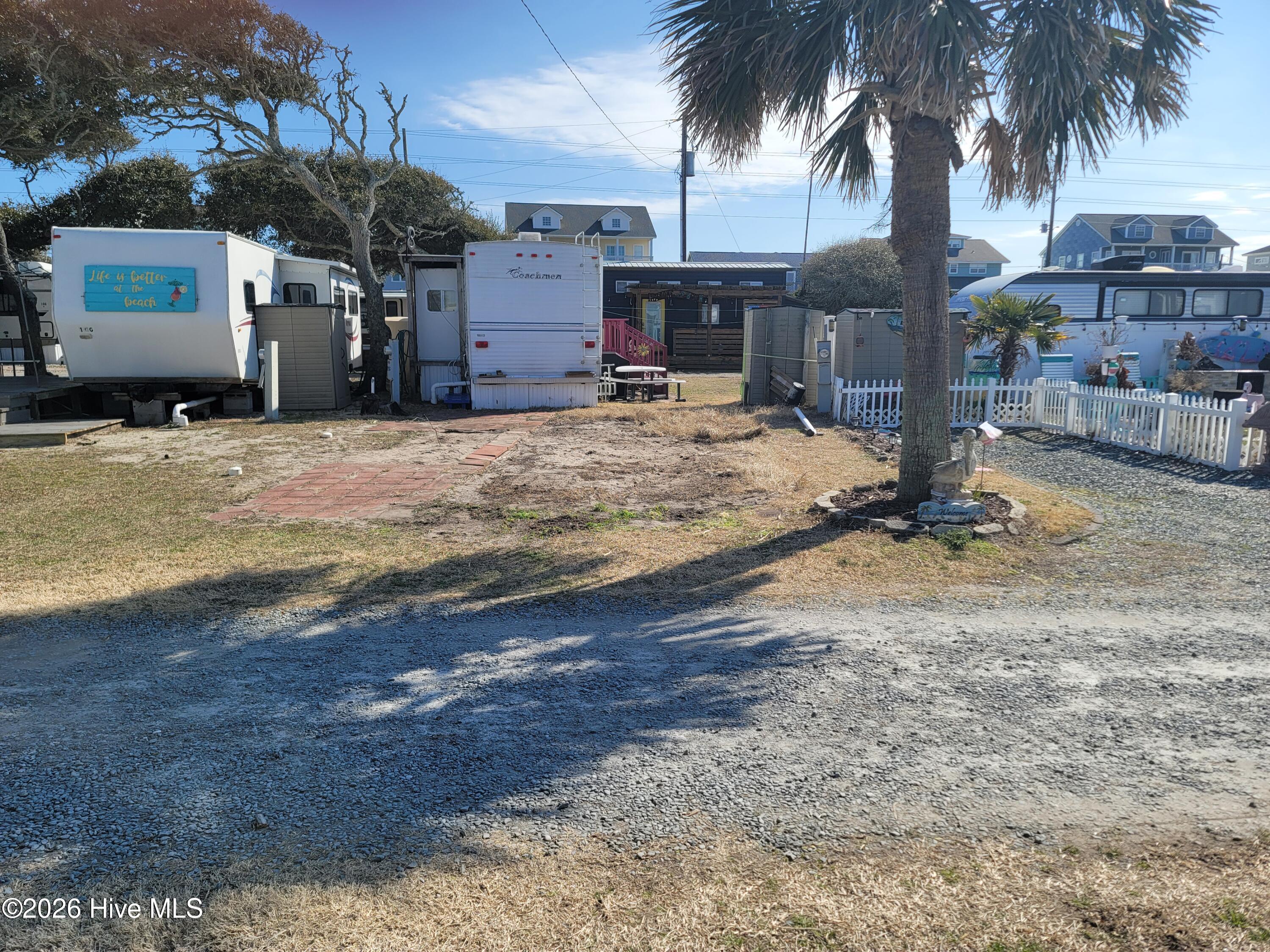 Gravel lot with several parked RVs, small sheds, a white fence, and a palm tree.