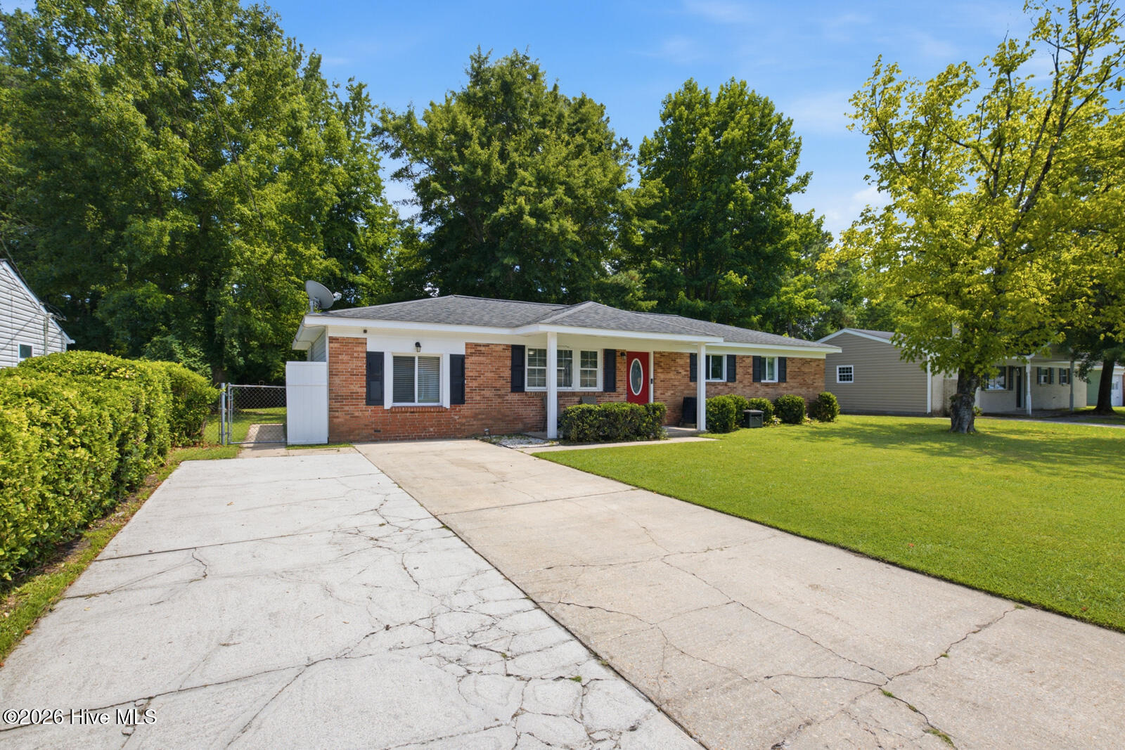 Brick ranch home with a red front door, broad driveway, green lawn, and mature trees.