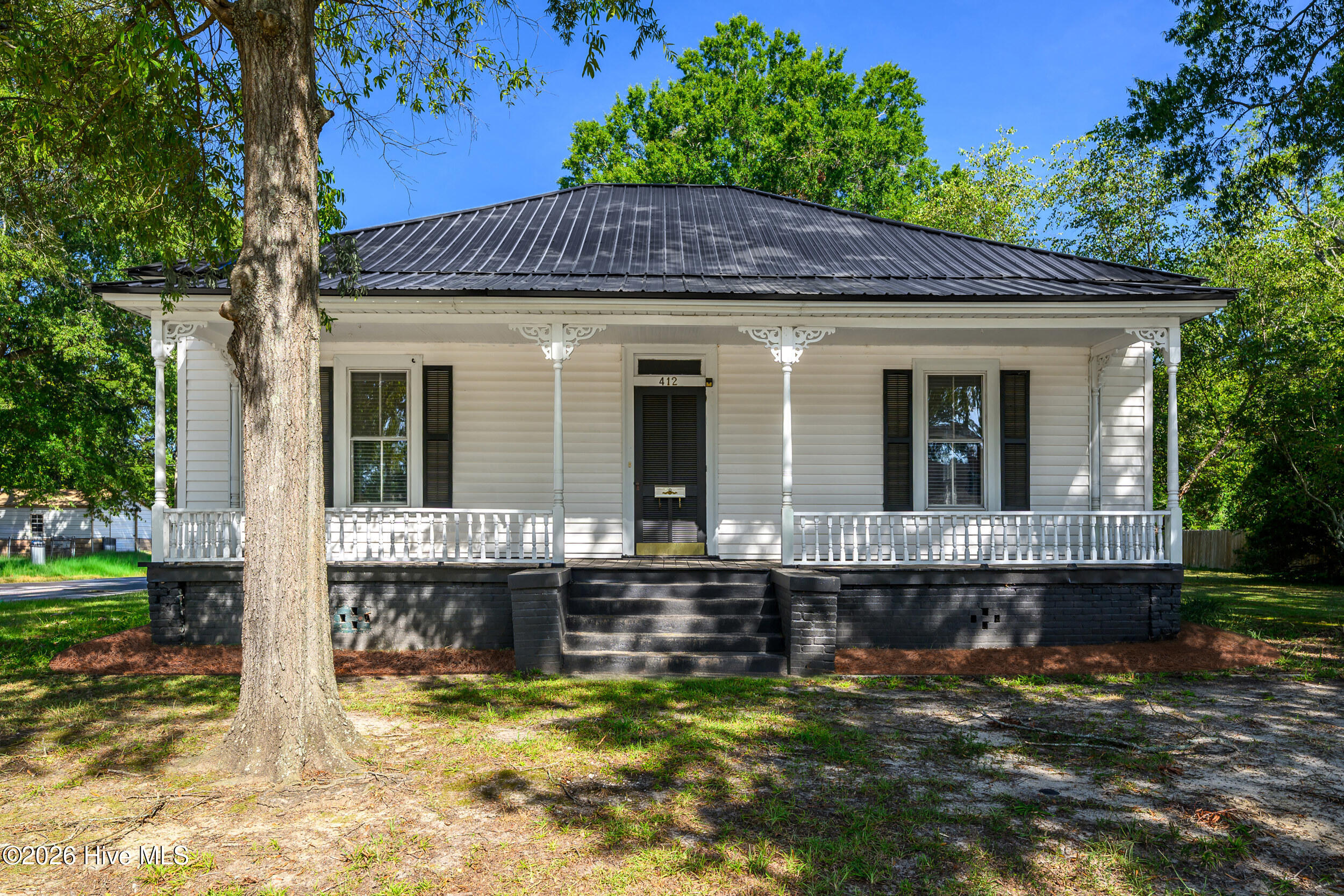 White house with a broad covered porch, black metal roof, and large tree in the front yard.