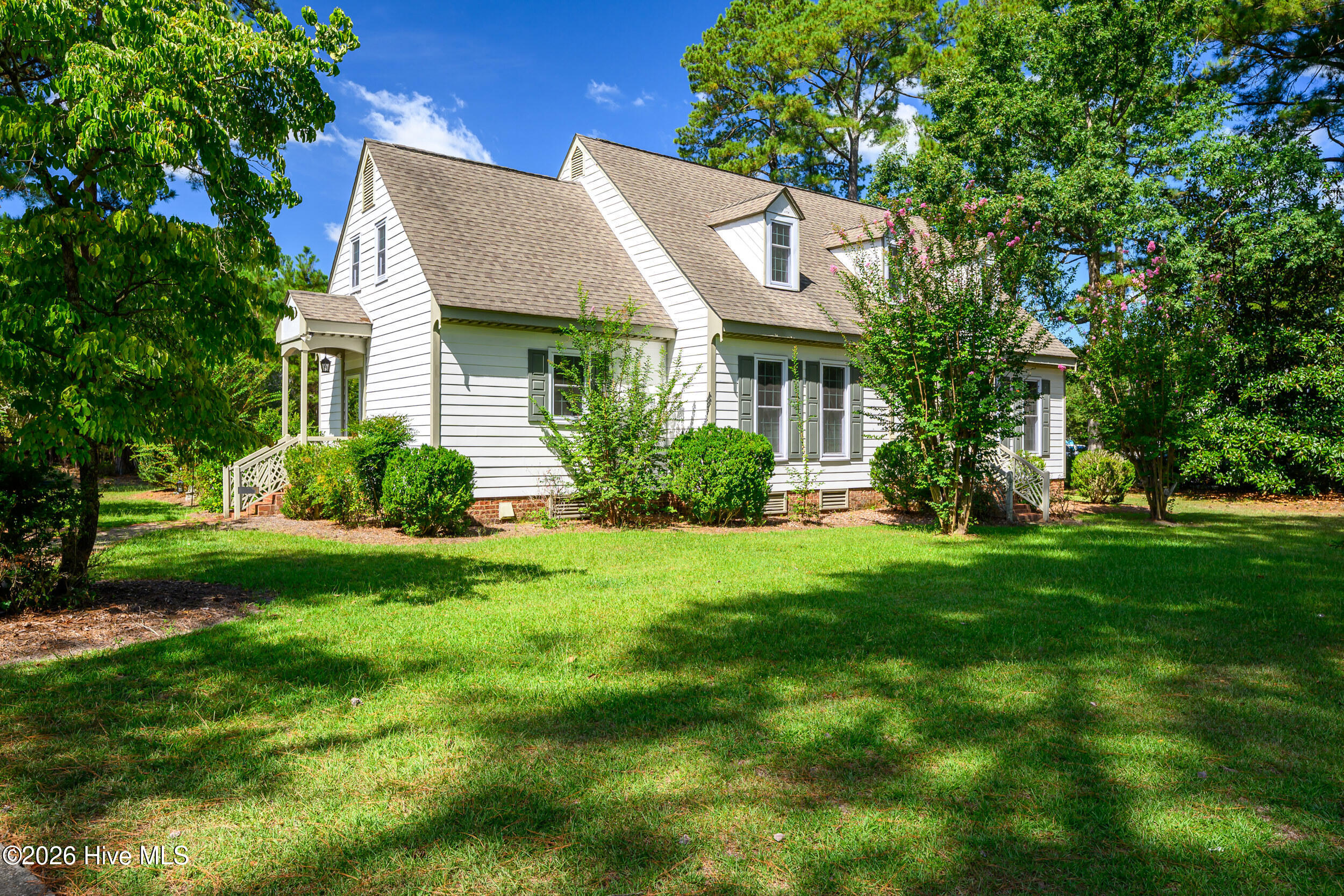 White-sided house with a steep gabled roof and dormers, surrounded by flowering shrubs and a shaded lawn.