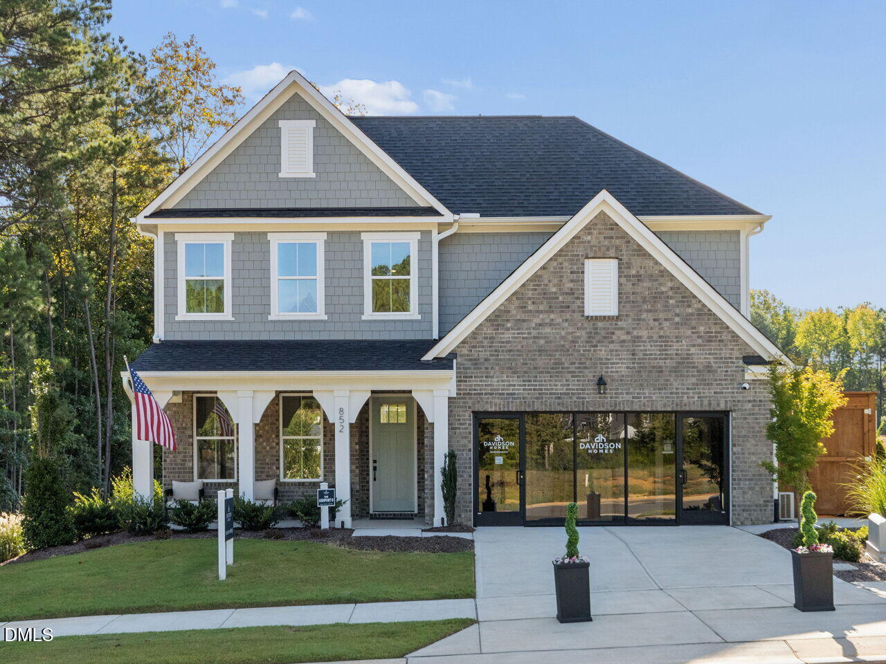 Two-story gray shingle and brick house with a covered porch, American flag, and wide driveway.