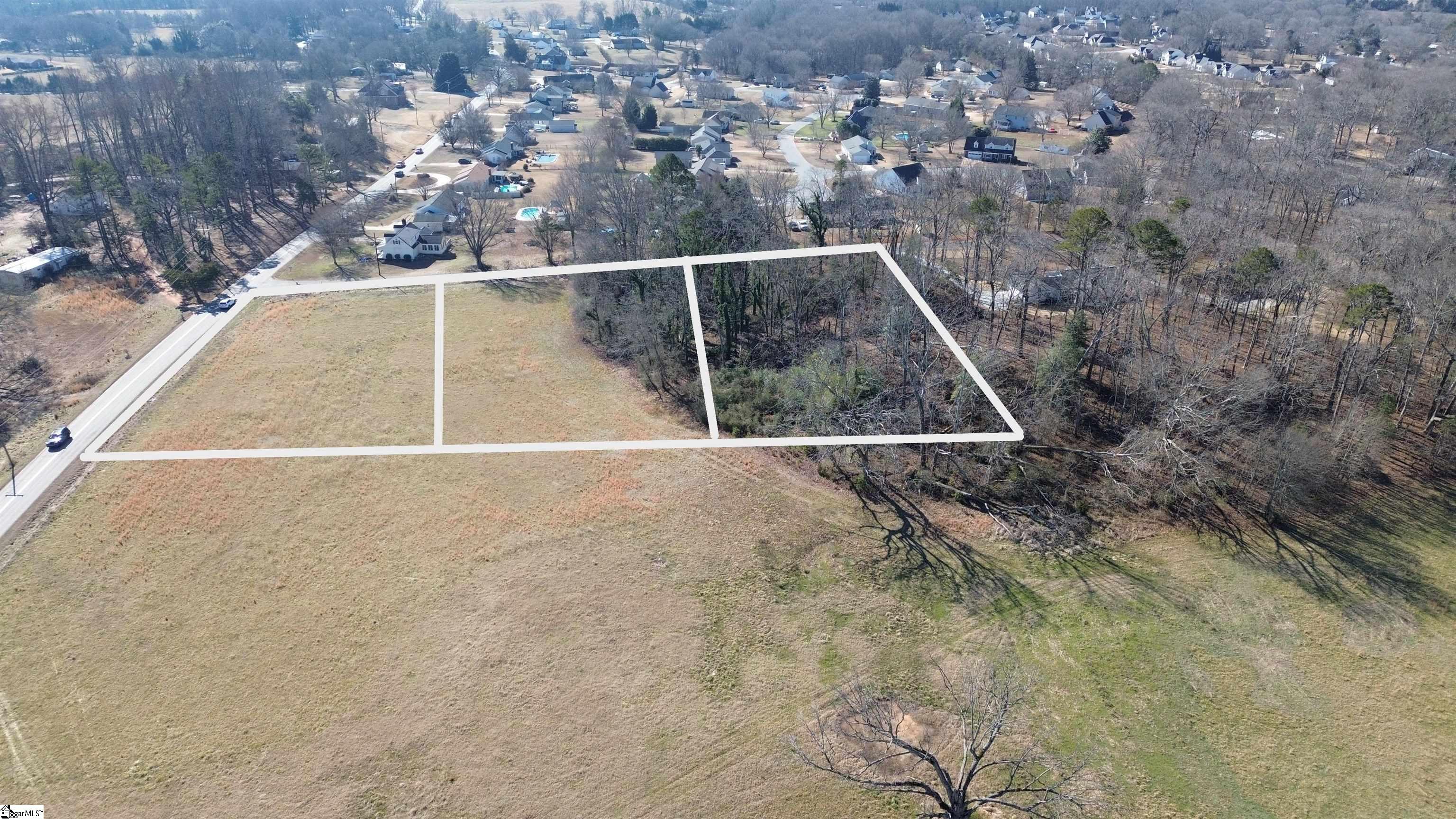 Aerial view of a vacant parcel outlined in white, with open grass, woods, and a road beside it.