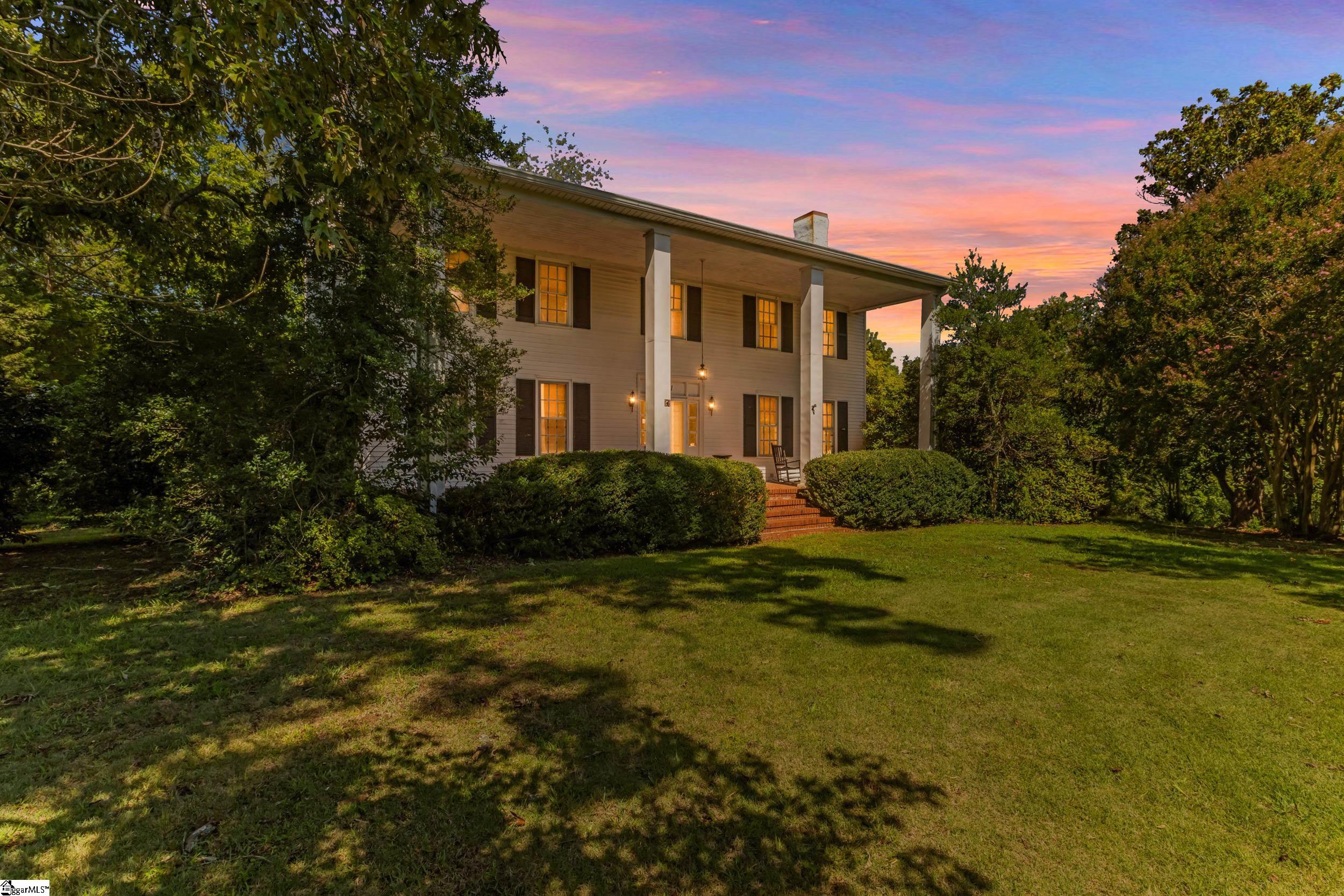 Two-story home with a columned porch and glowing windows at dusk, framed by mature trees.