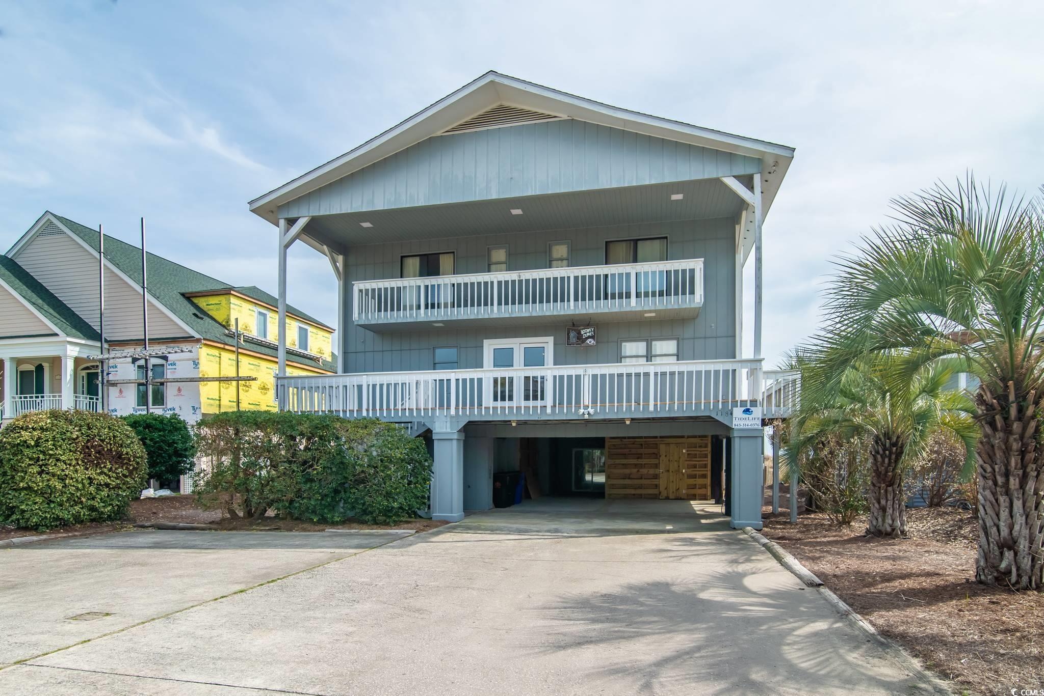 Blue-gray raised house with two white-railed balconies, an open carport, and palm trees beside the driveway.