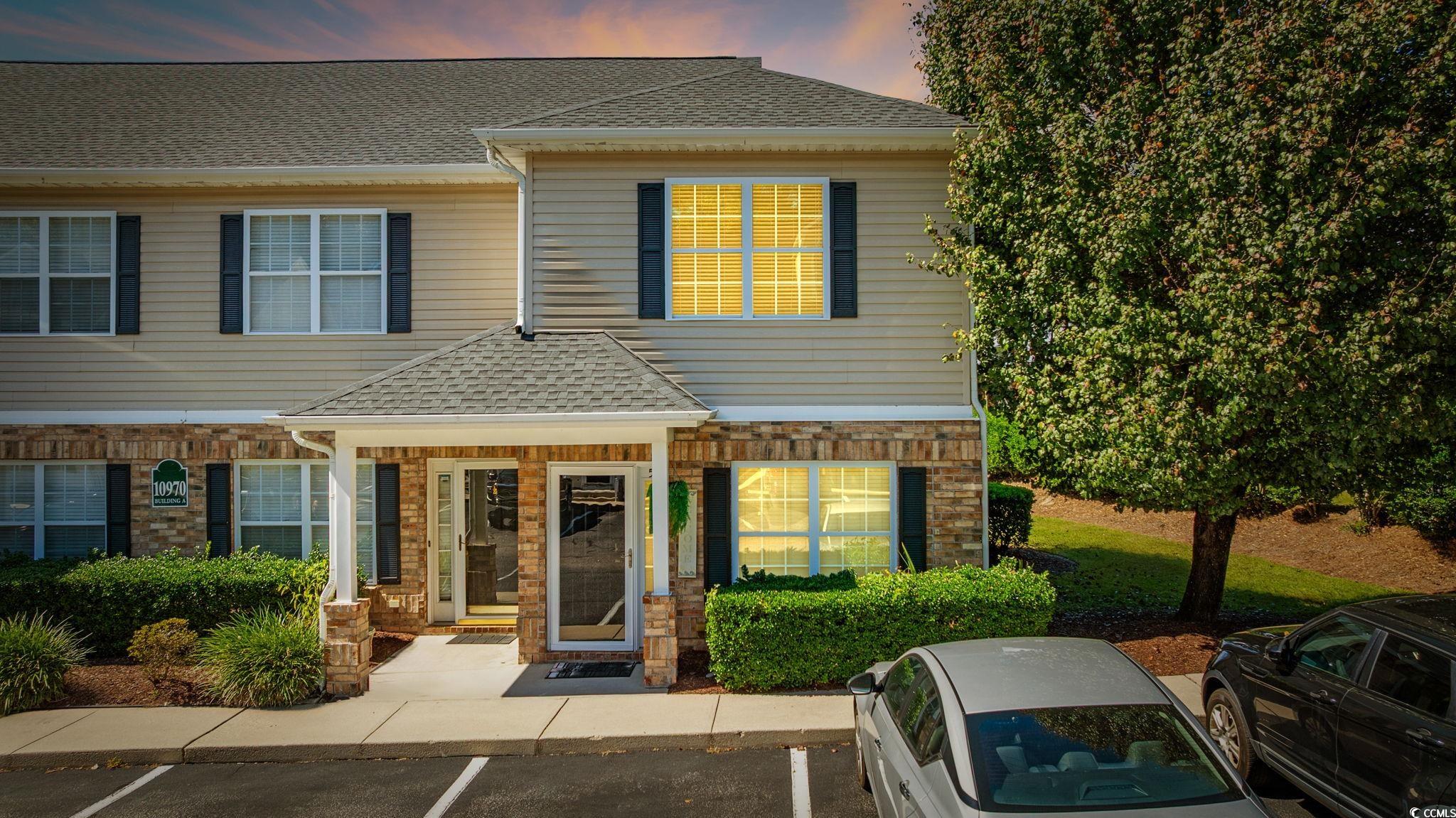 Two-story building with a brick entrance, tan siding, and parked cars in front.