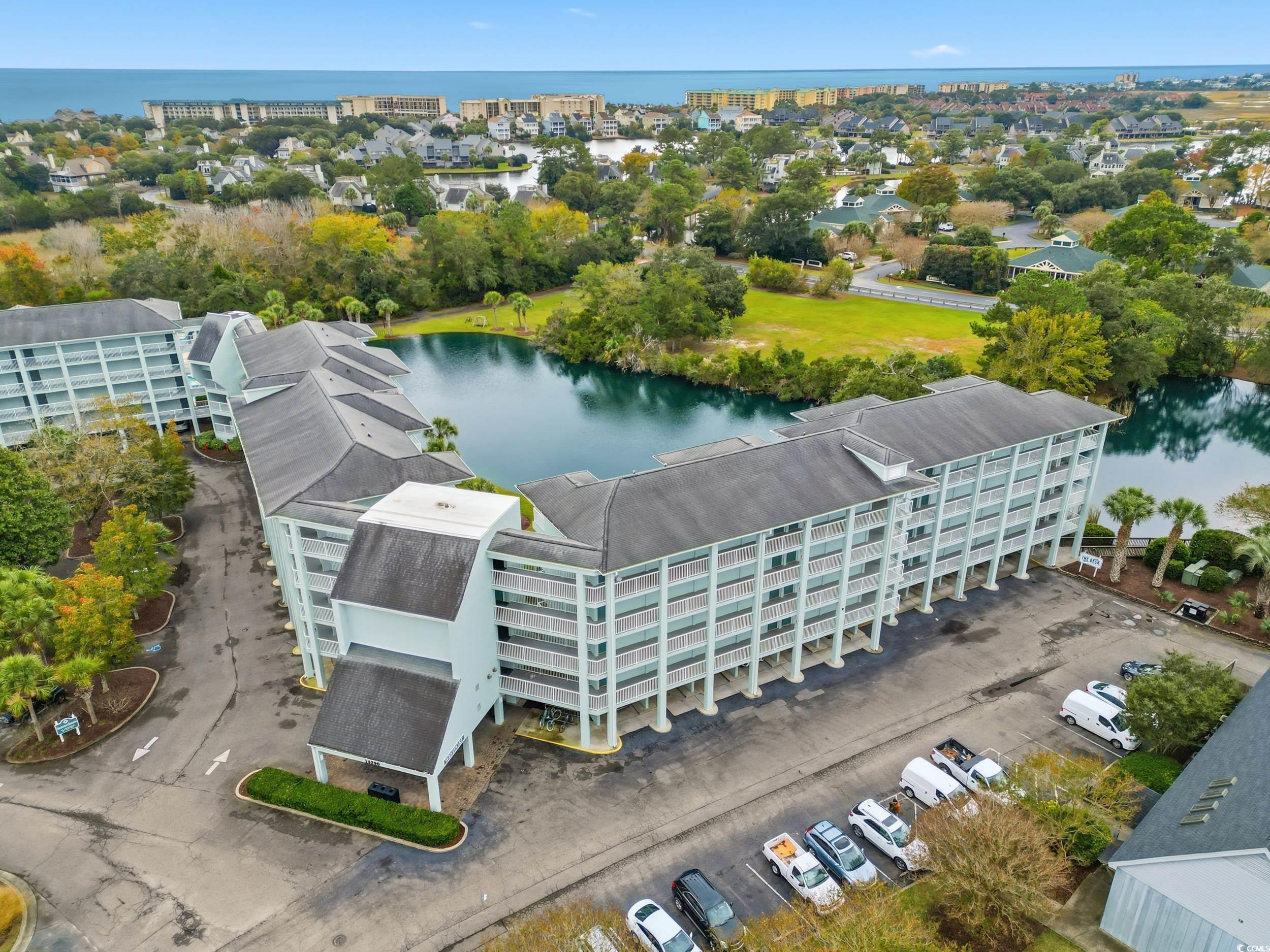 Aerial view of a condominium complex beside a lake, with the ocean and coastal homes beyond.