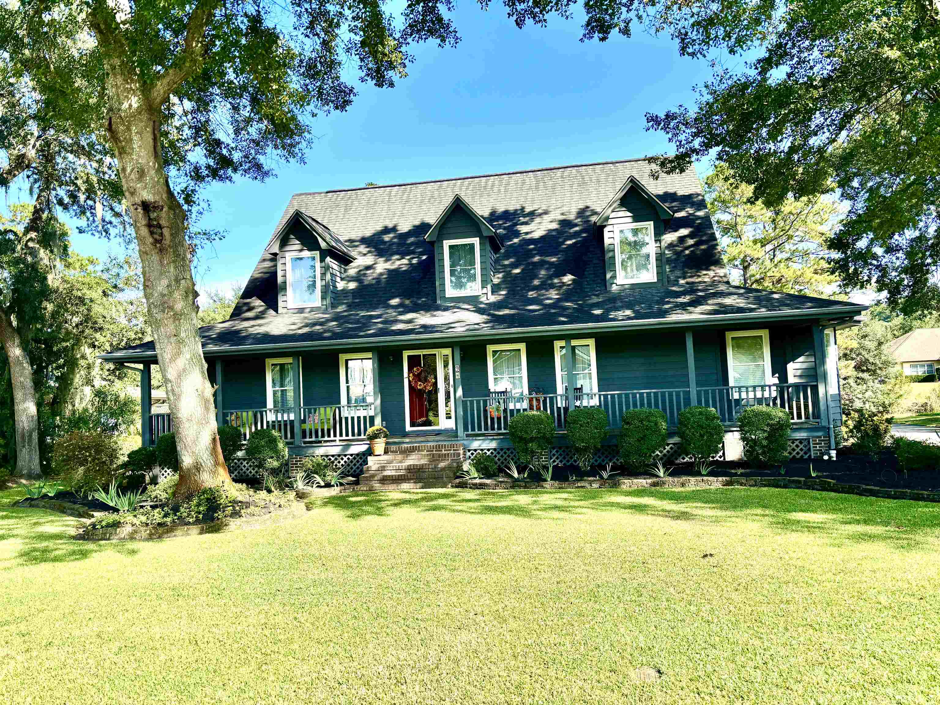 Dark teal house with three dormer windows, a covered porch, and a broad lawn beneath mature trees.