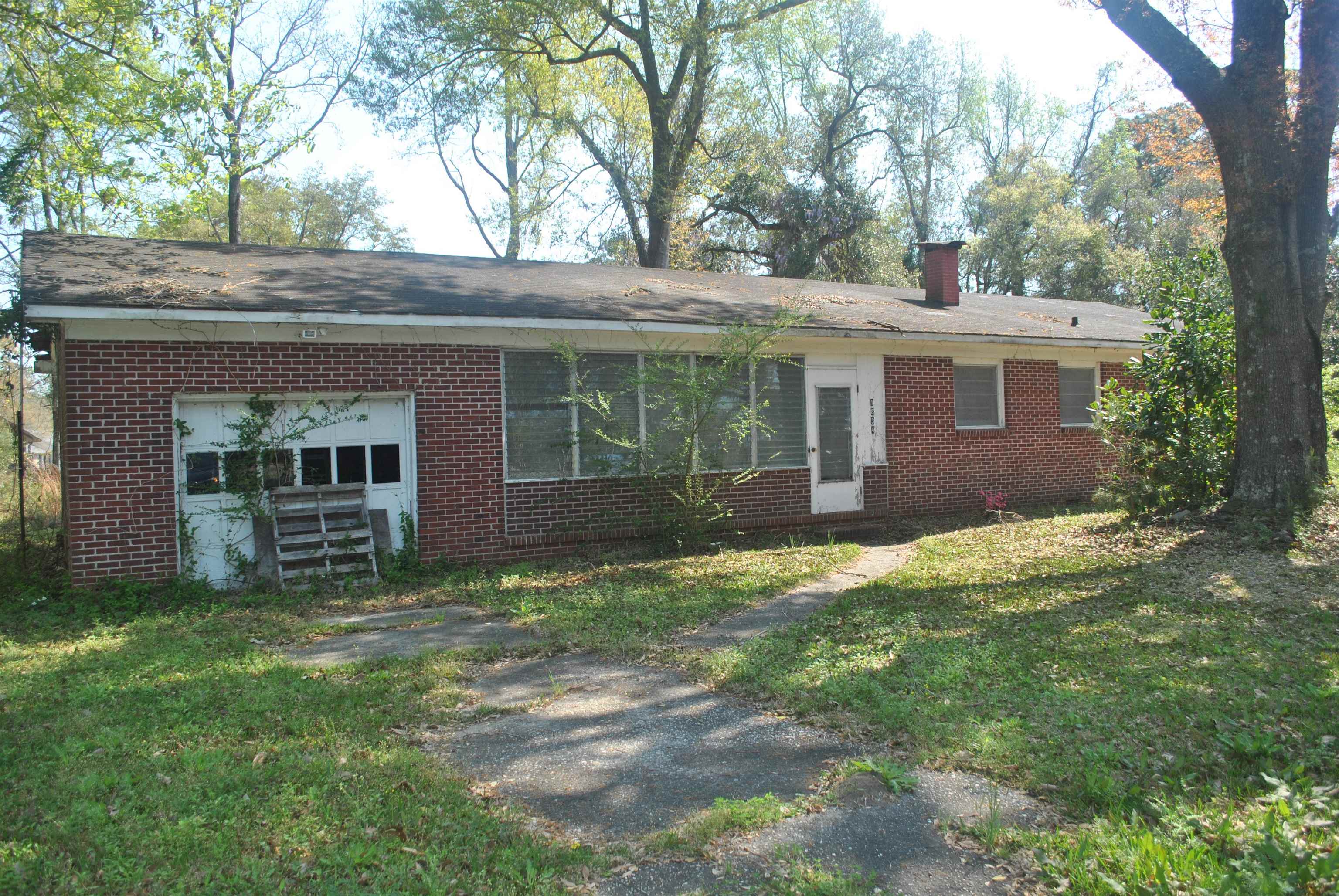 Single-story brick house with a low roof, mature trees, and a cracked walkway through an overgrown yard.