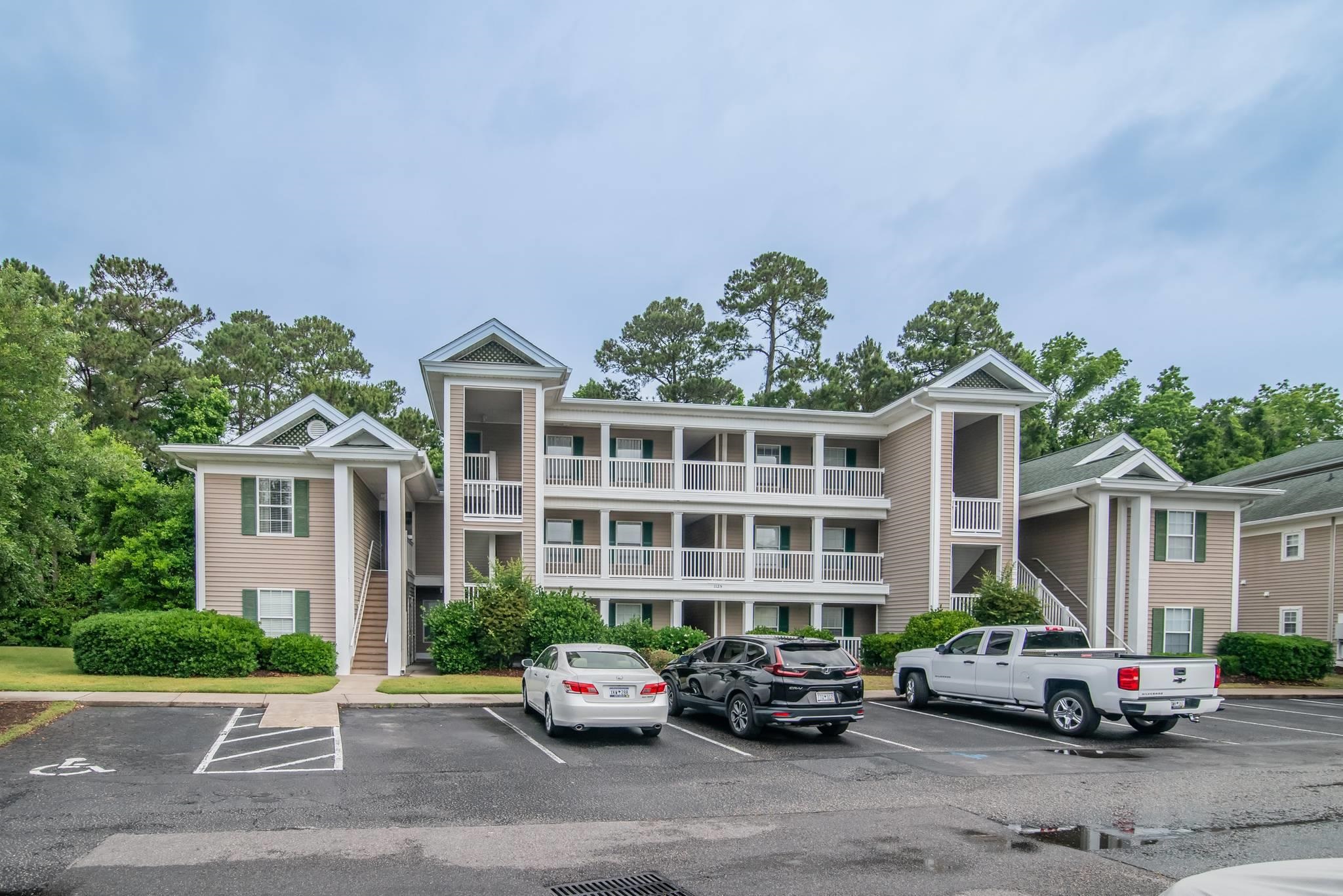 Three-story beige condominium building with long white balconies and cars parked in front.