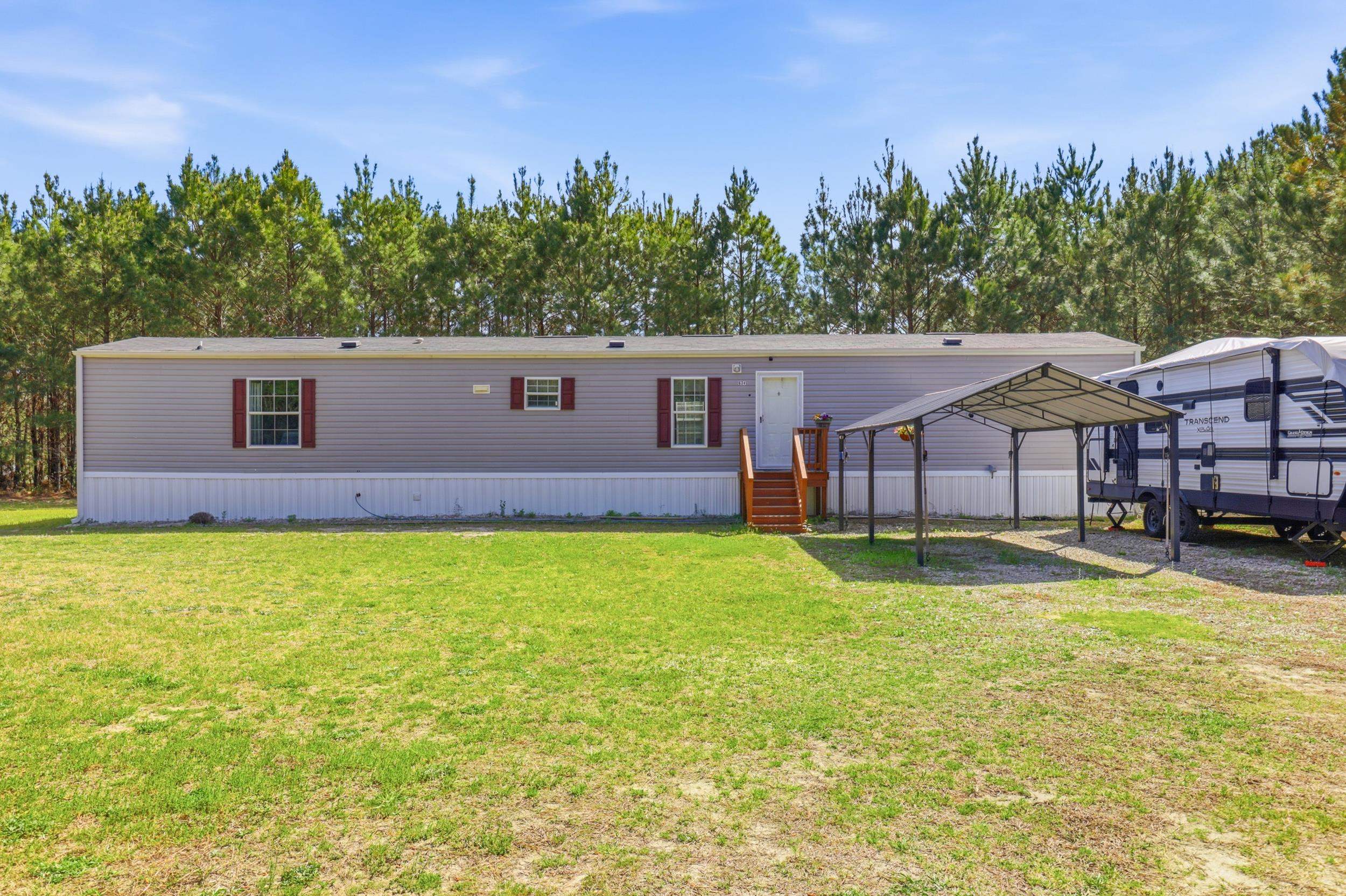 Gray manufactured home with burgundy shutters, wooden steps, and a carport beside a travel trailer.