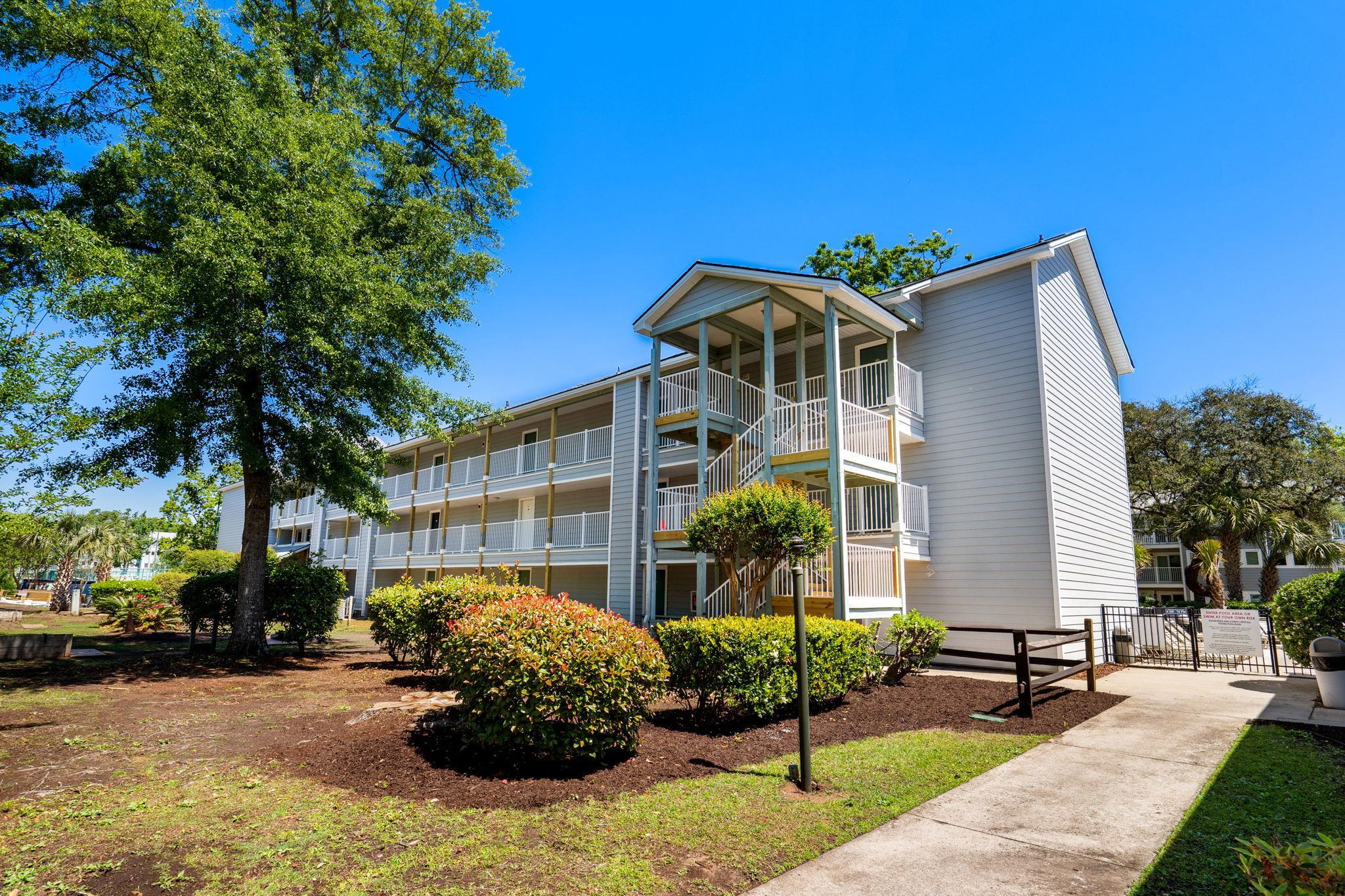 Three-story gray condominium building with white balconies, an exterior staircase, and landscaped grounds.