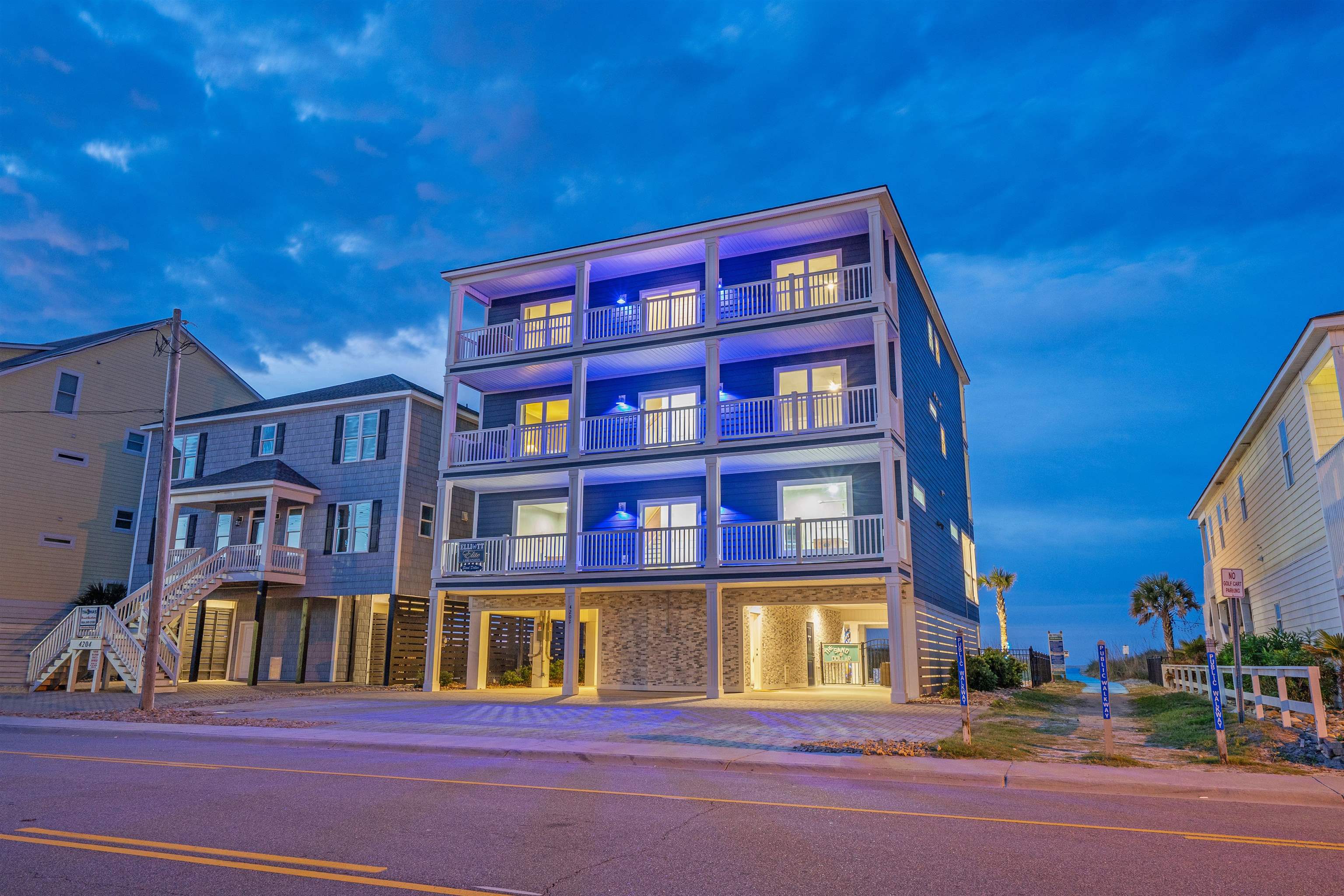 Three-story coastal home with stacked balconies glowing blue at dusk beside a beach access walkway.