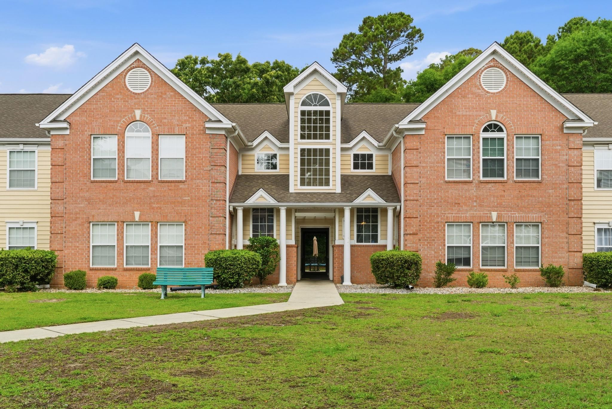 Brick apartment building with a columned central entrance, turquoise bench, and broad front lawn.
