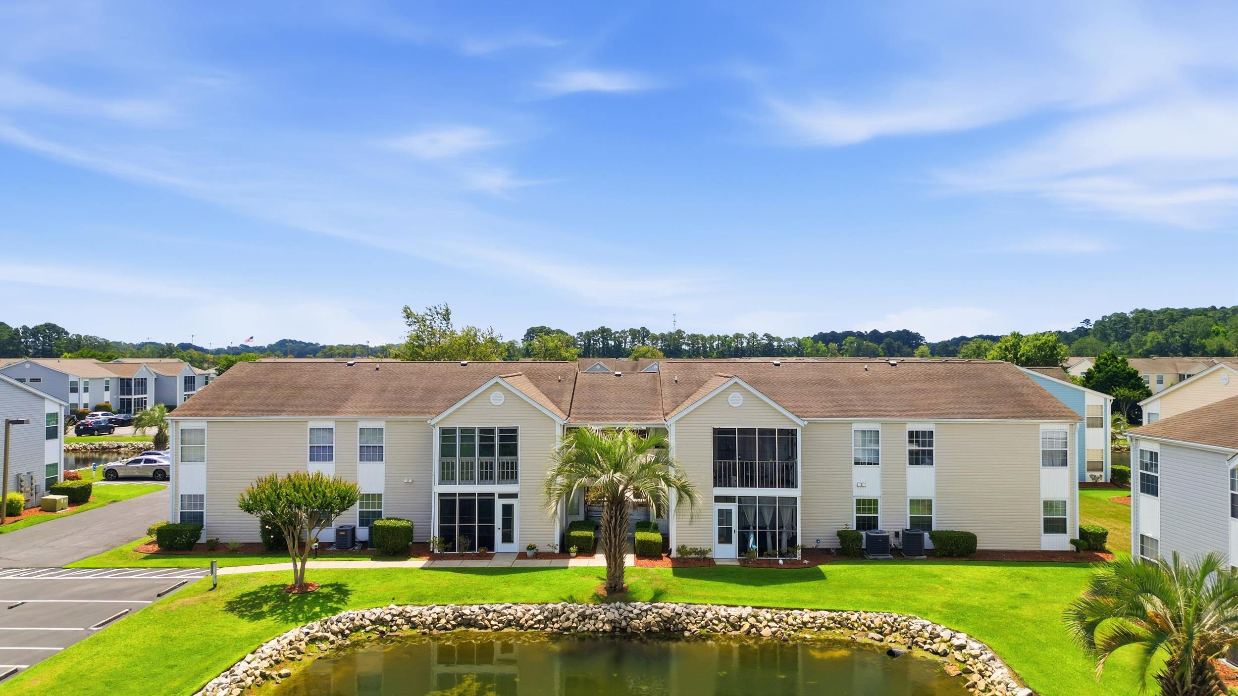 Aerial view of a beige apartment building beside a rock-edged pond and landscaped lawn.