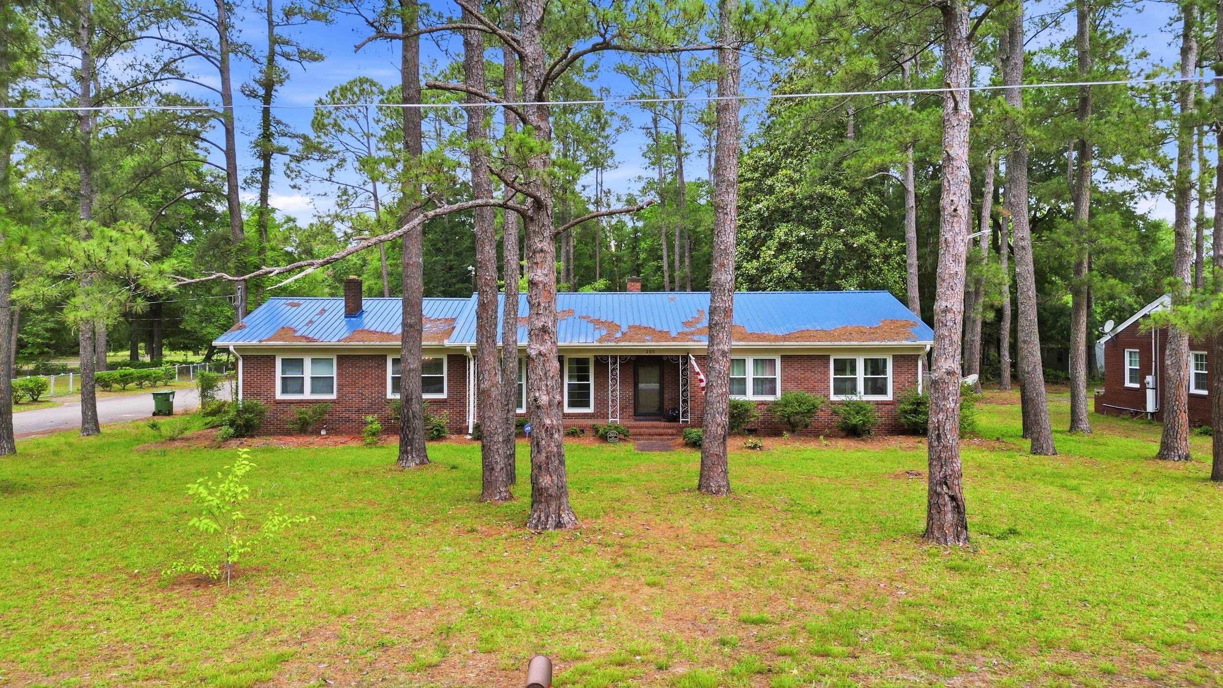Red-brick ranch home with a blue metal roof among tall pine trees.