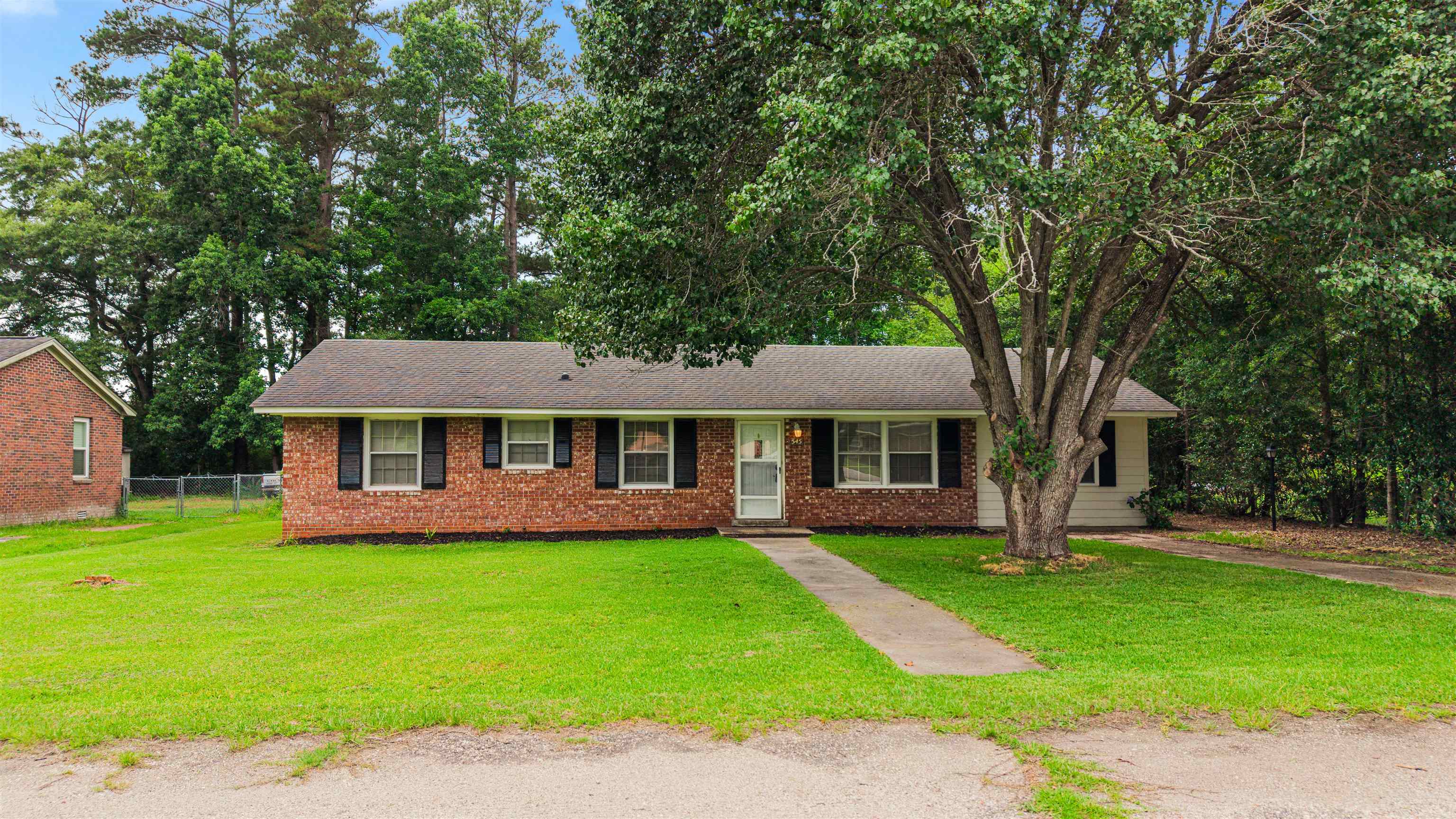 Single-story brick ranch home with black shutters, a broad lawn, and a large tree in front.