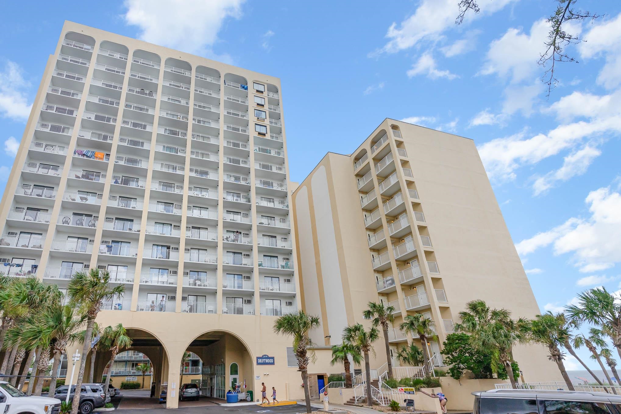 Driftwood beachfront condominium with stacked white balconies, palm trees, and a glimpse of ocean.