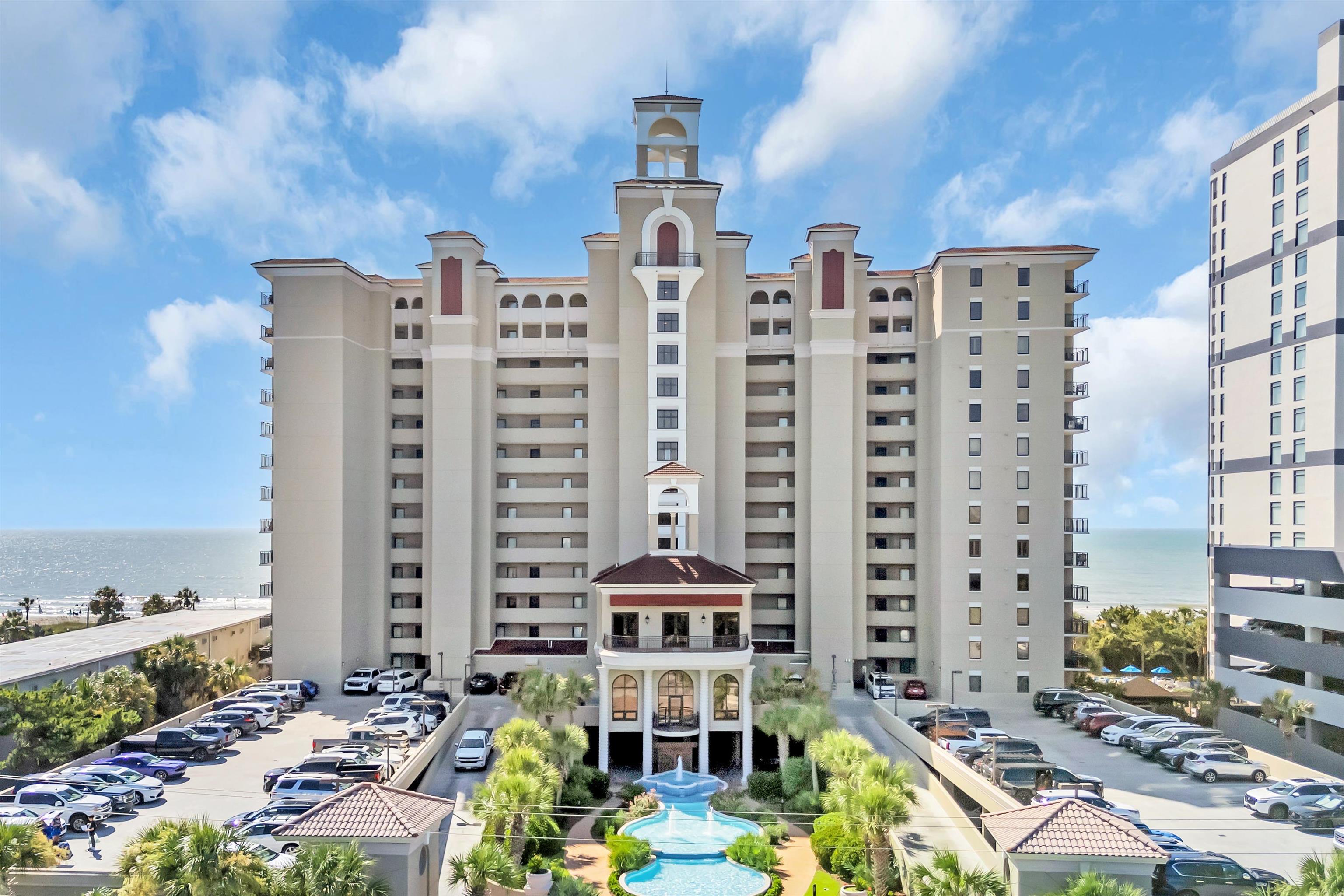 Oceanfront condominium tower with a landscaped pool courtyard and parking, viewed from above.