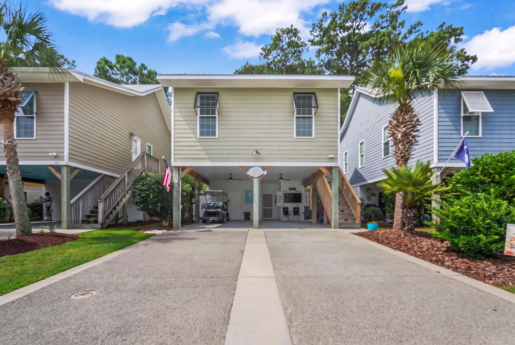 Raised light-green home with a covered carport, broad driveway, exterior stairs, and palm trees.