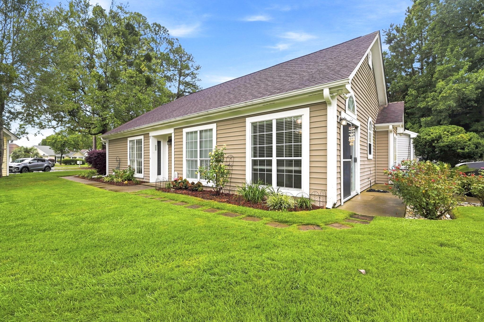Beige house with a steep roof, large windows, landscaped beds, and a broad front lawn.