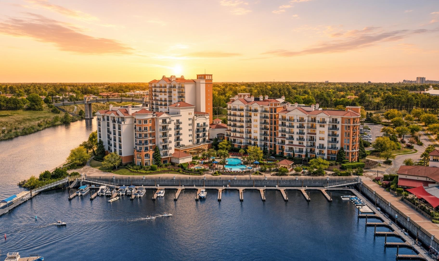 Waterfront condominium complex beside a marina, with boats, a pool, and a river at sunset.