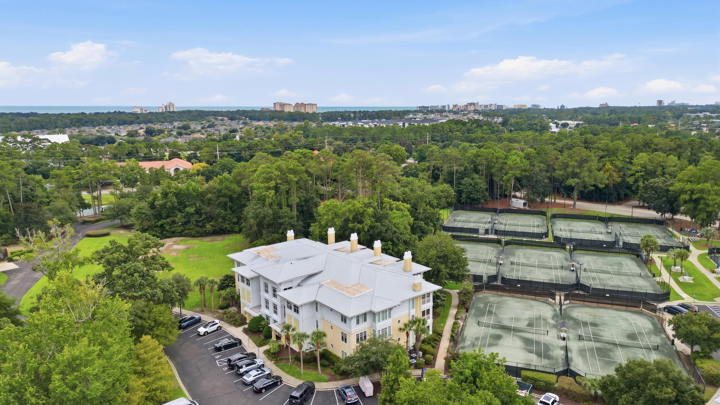 Condominium building beside tennis courts, with wooded grounds and the ocean visible in the distance.