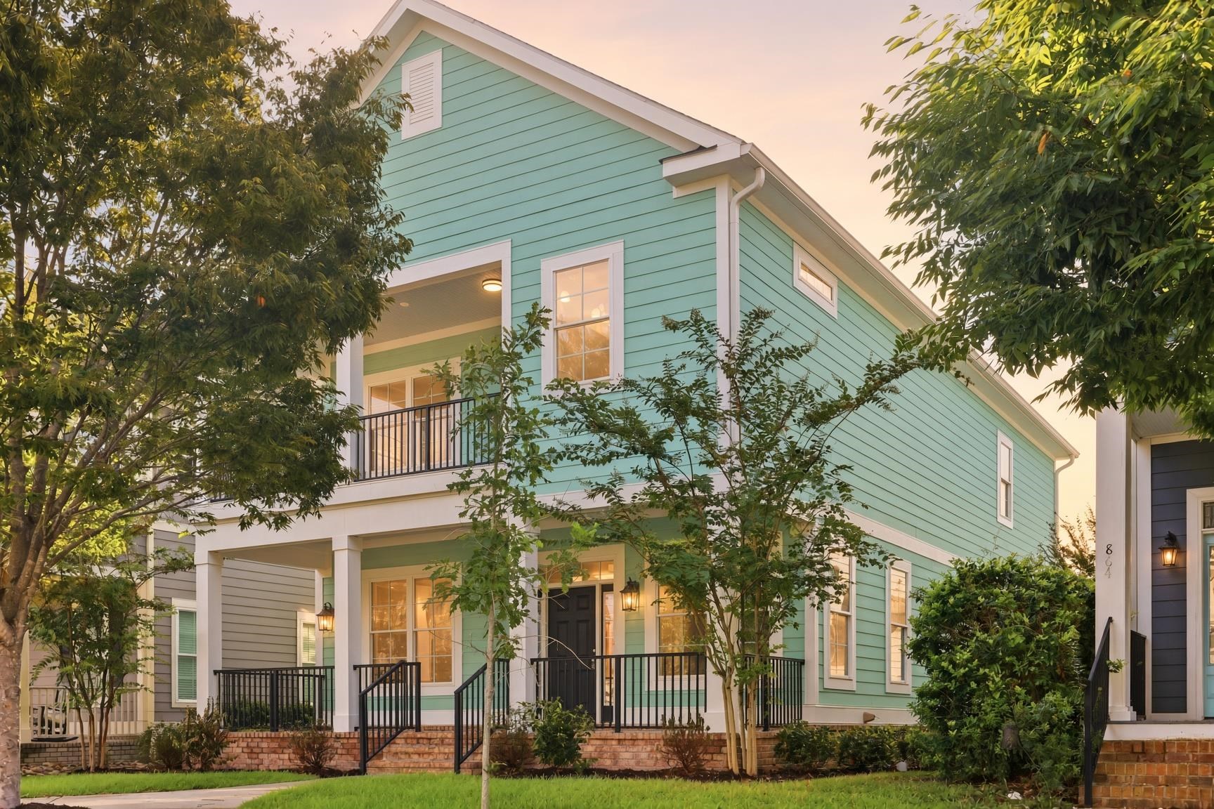 Aqua two-story house with a covered porch, black railings, brick steps, and leafy trees in warm evening light.