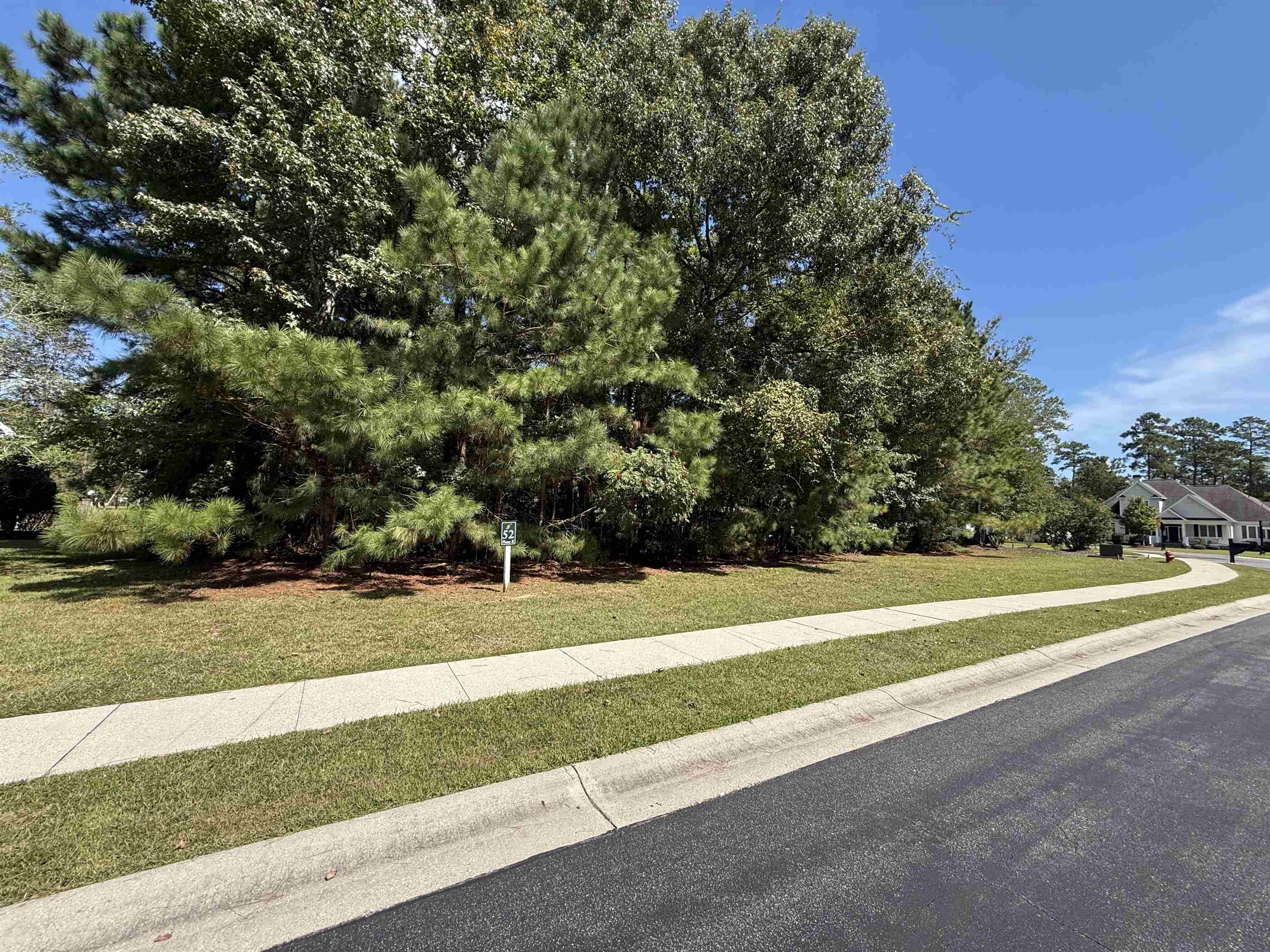 Wooded residential lot with mature trees, grassy frontage, and sidewalk along the street.