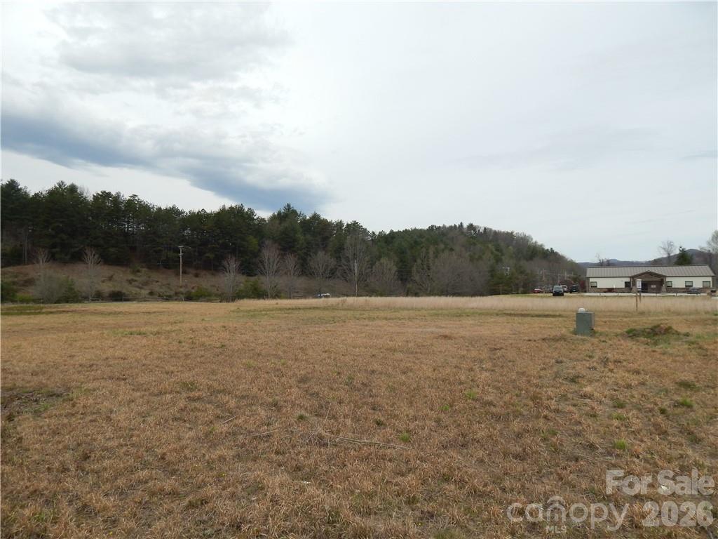 Vacant grassy lot with wooded hills behind it and a small building to the right.