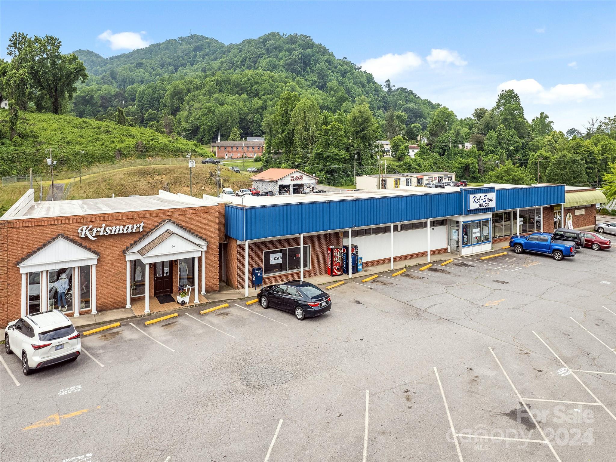 Commercial storefronts and parking lot below wooded hills, including a brick shop and blue-fronted drugstore.