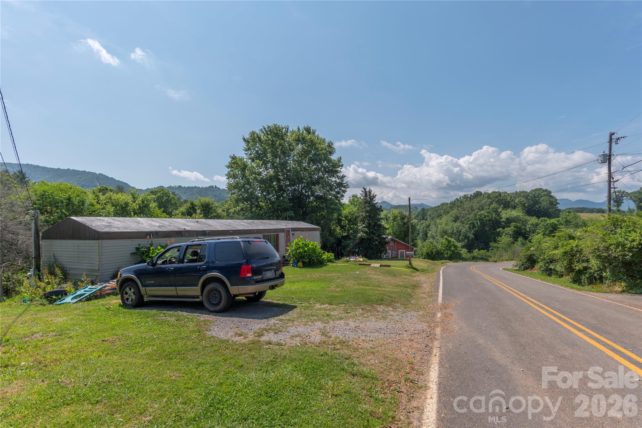 Roadside property with a long metal-sided building, parked SUV, and wooded mountain hills beyond.