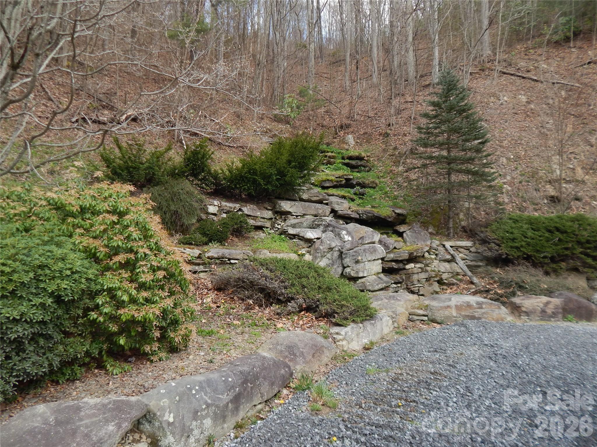Wooded hillside with layered stone retaining walls beside a gravel drive.