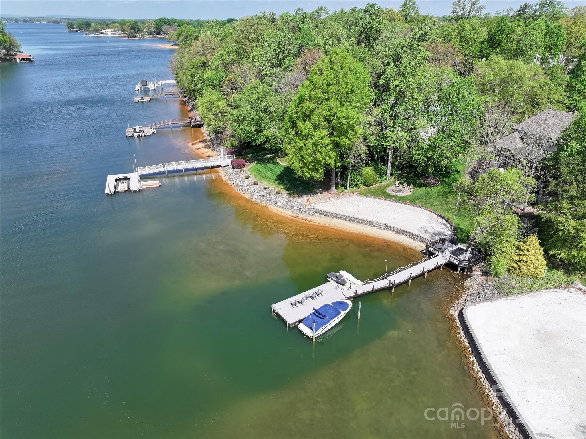 Aerial view of a wooded lakefront property with multiple docks, a moored boat, and a sandy beach.