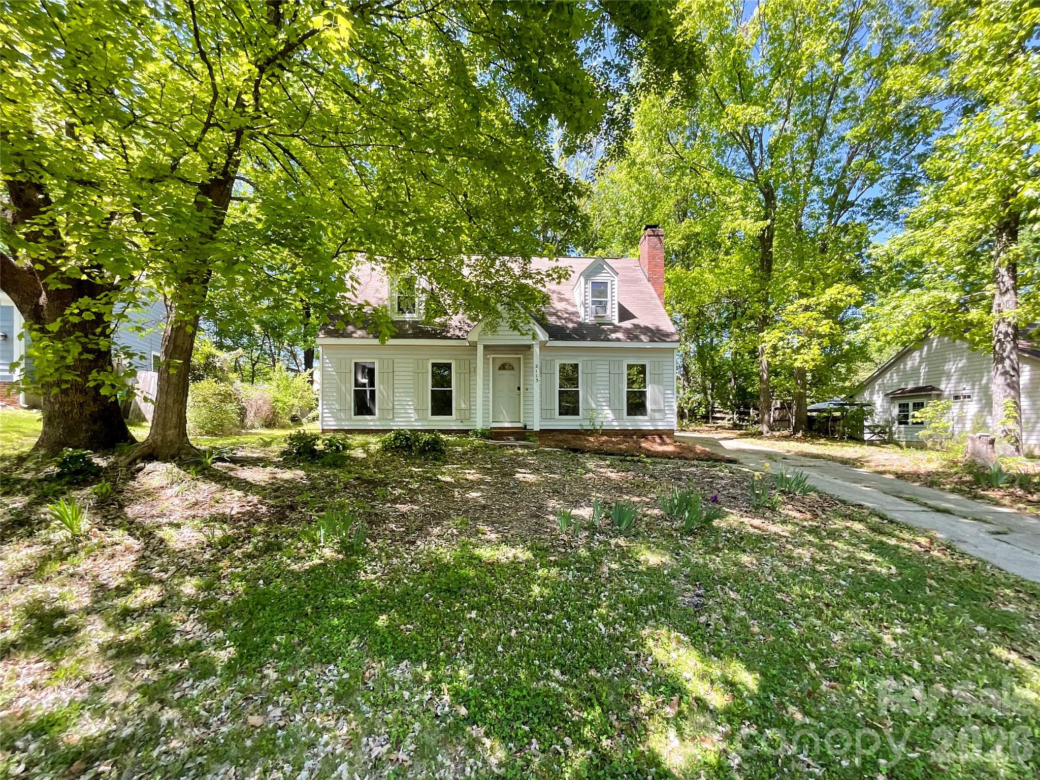 White Cape Cod-style house with a brick chimney, framed by mature trees and a shaded front yard.
