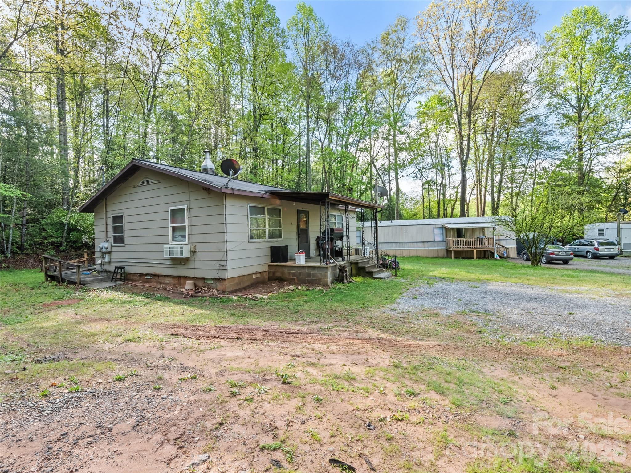 Small light-sided home with a covered porch on a wooded lot beside a manufactured home.