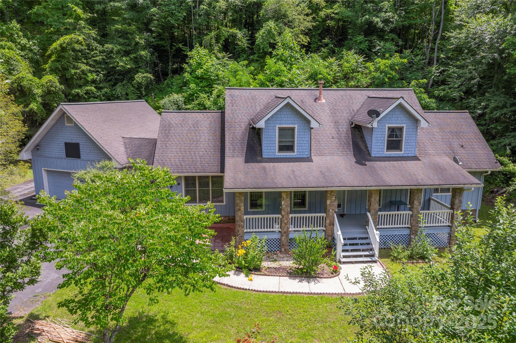 Blue-sided home with a stone-columned porch, attached garage, and wooded surroundings.