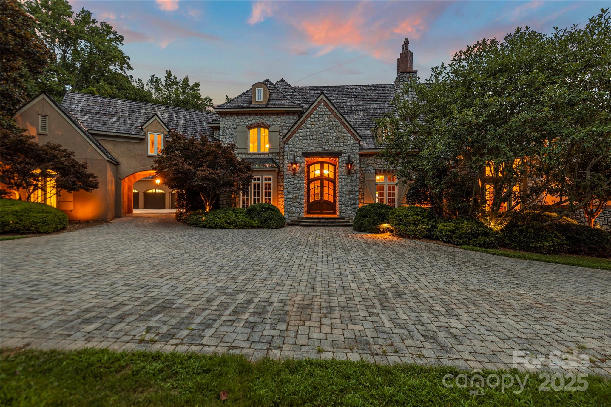 Stone house with a warmly lit arched entrance and broad paver driveway at dusk.