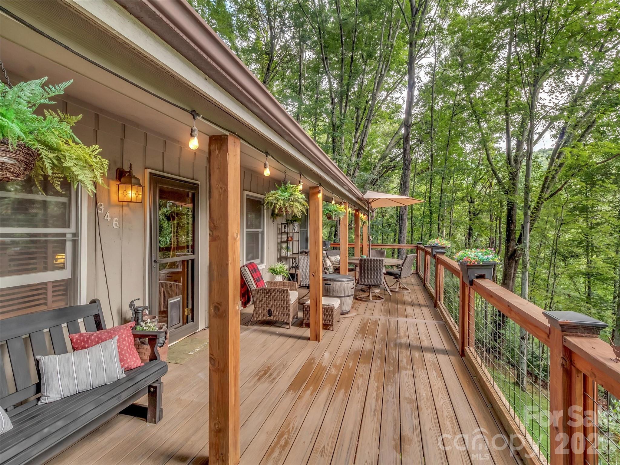 Furnished wooden deck overlooking trees, with wicker seating, string lights, and a wood-and-wire railing.